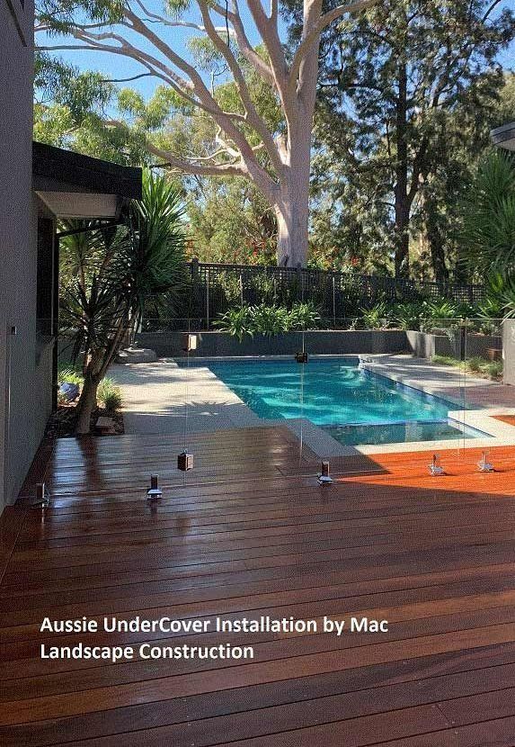 Wooden deck beside a pool. Clear glass fencing. Tree in the background. Bright sunny day.