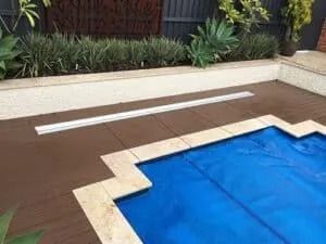 Pool covered with blue tarp, surrounded by brown deck, beige coping, and landscaping.