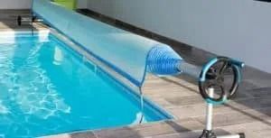 A swimming pool with blue water covered by a blue, rolled-up cover. A hand crank is visible.