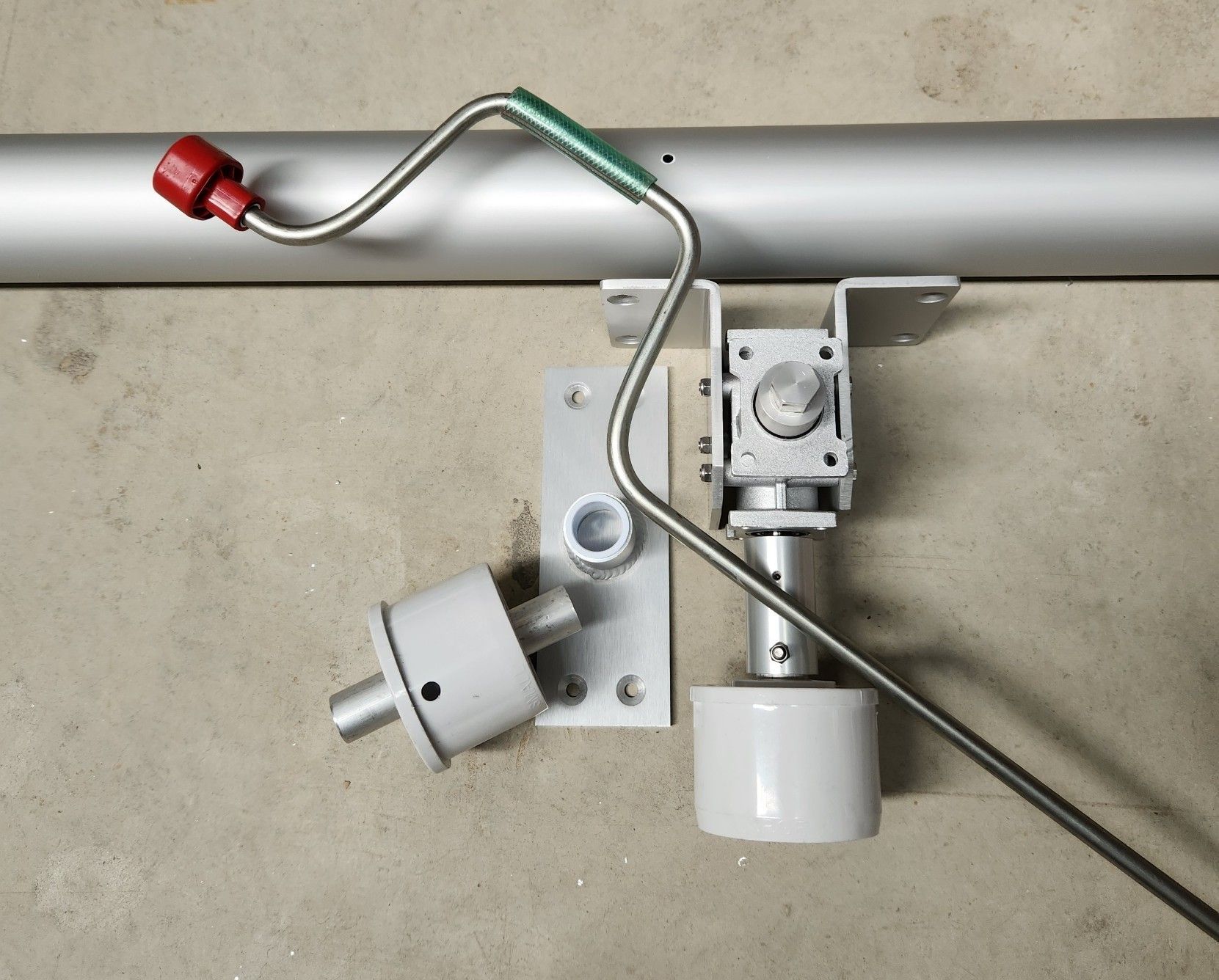Silver manual pool cover mechanism parts on a gray surface, including a pole, bracket, and handle.