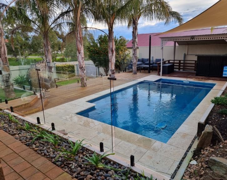 Rectangular pool with blue water and light stone surround, next to a brick patio and landscaped yard.