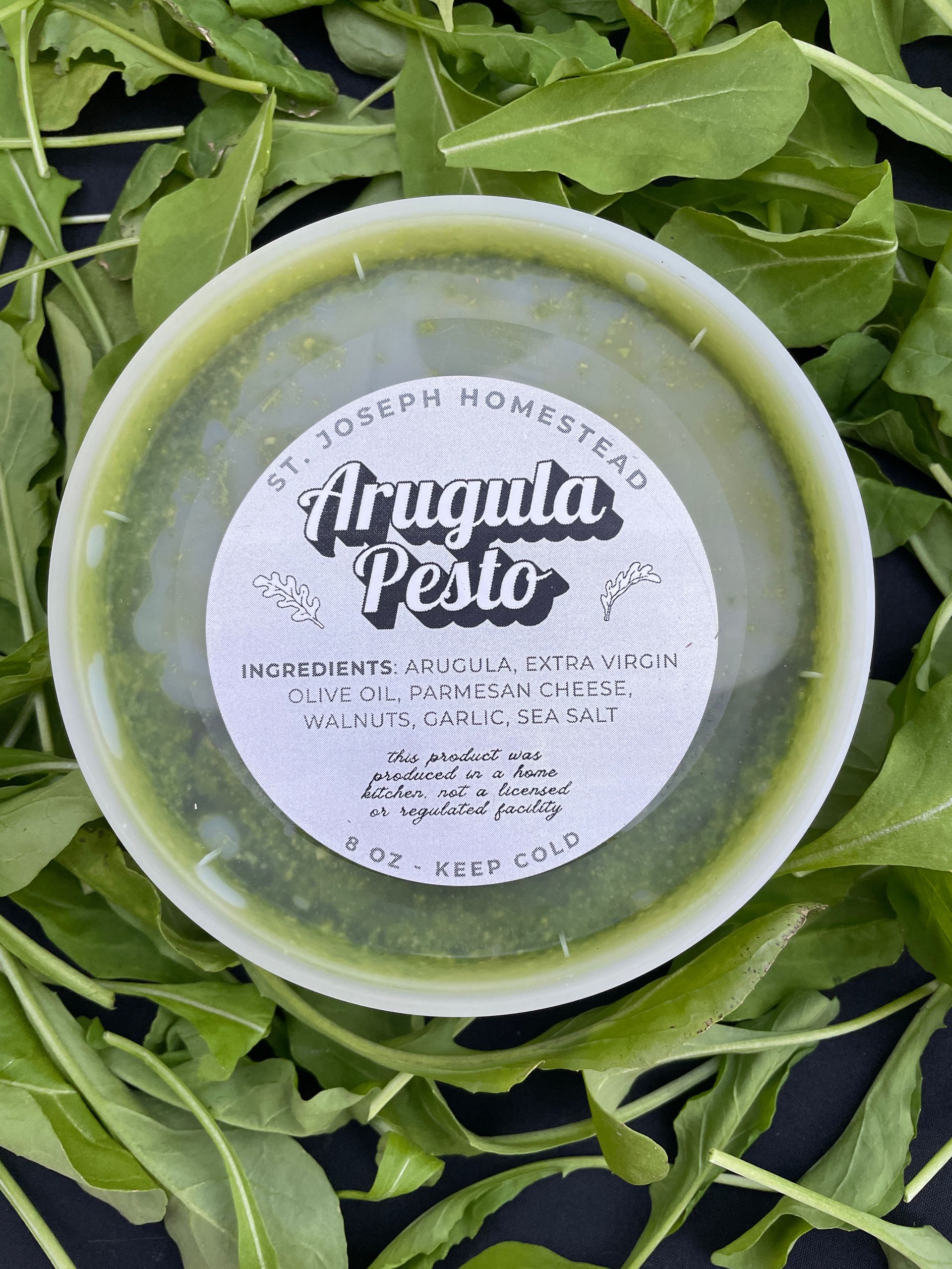 Green jar labeled “Arugula Pesto” sitting on fresh arugula leaves.