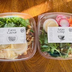 Two packaged salads on a wooden table: Farm Caesar and Asian Crisp, with visible greens and dressing cups