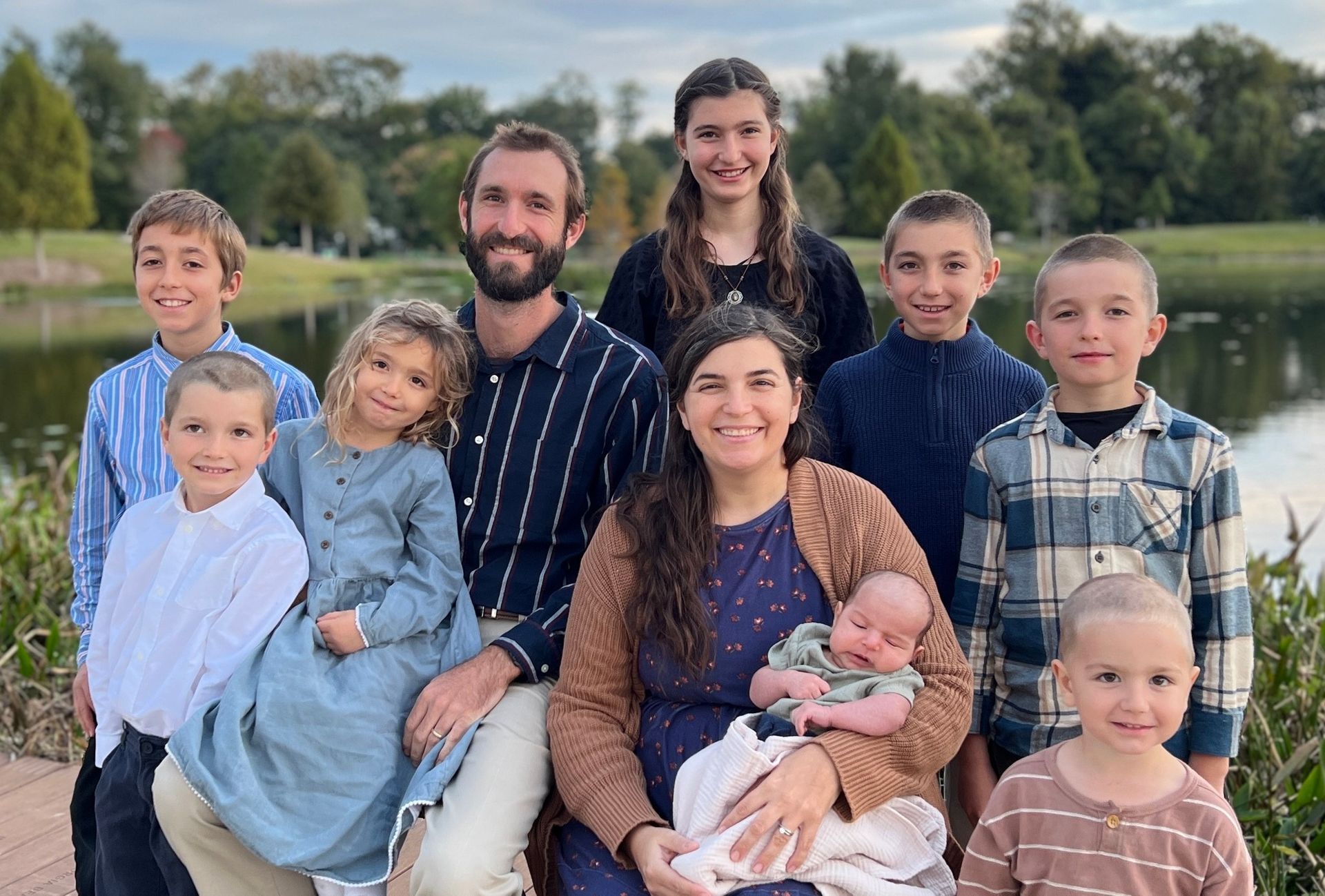 Large family posing outdoors by a lake, with parents holding a baby and children gathered around them.