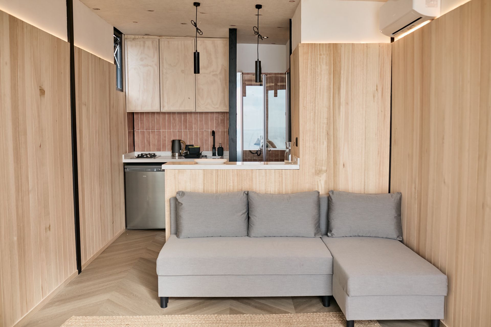 Modern studio apartment with light wood finishes, gray sectional sofa, and compact kitchen in the background
