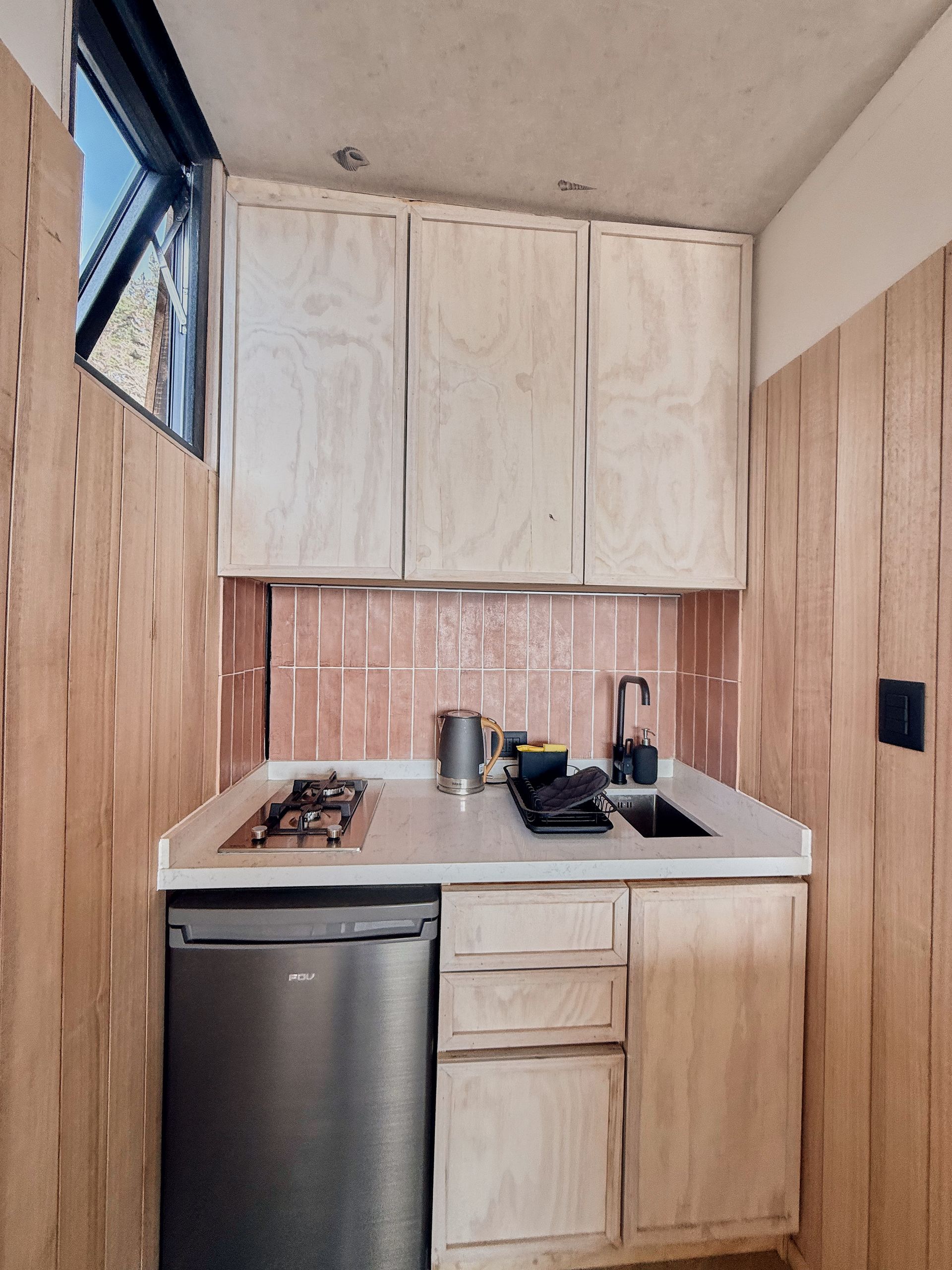 Compact modern kitchenette with light wood cabinets, sink, stove, mini fridge, and white countertop.