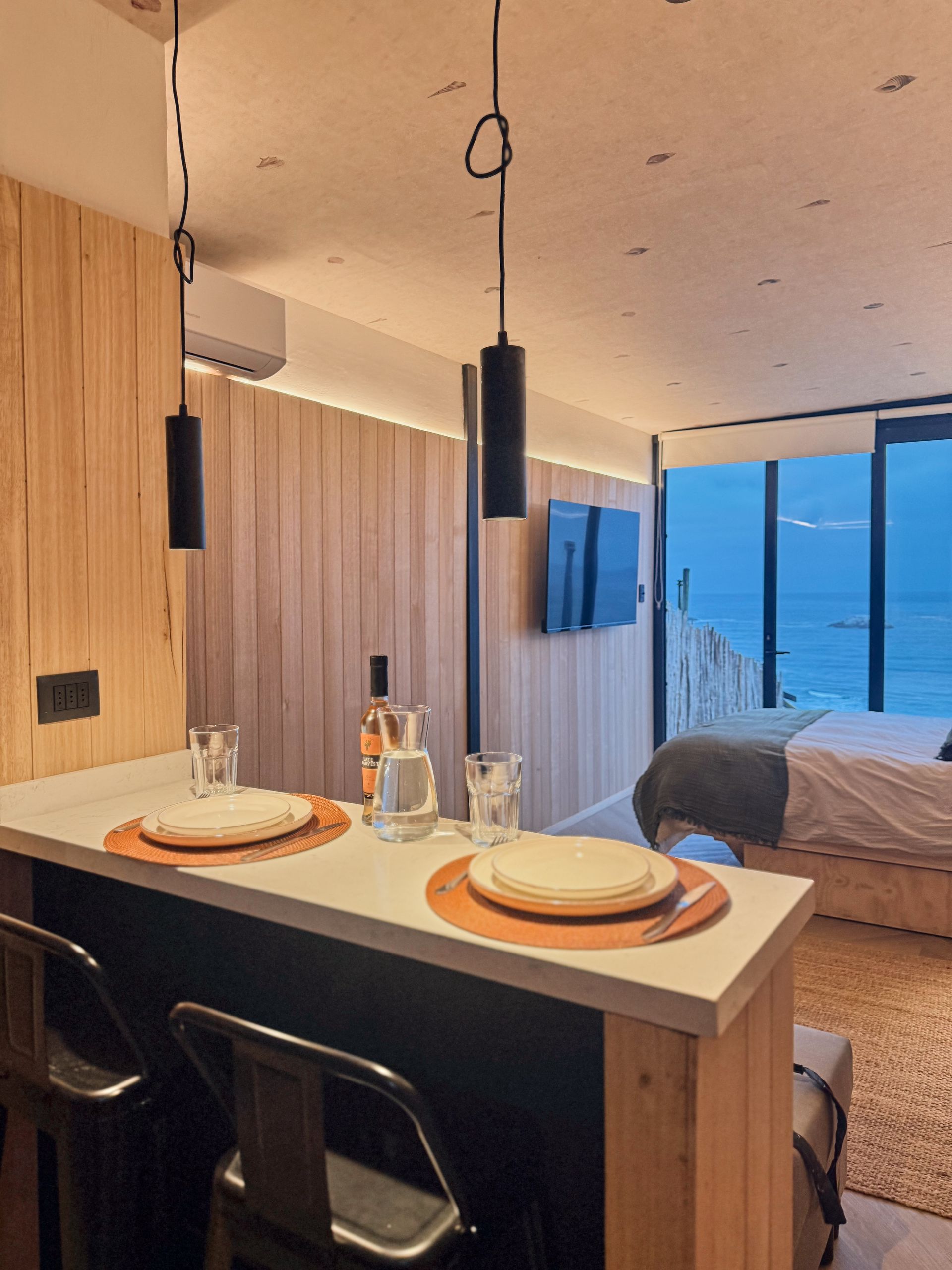 Modern apartment kitchen with dining table, pendant lights, bed, and ocean view through large windows