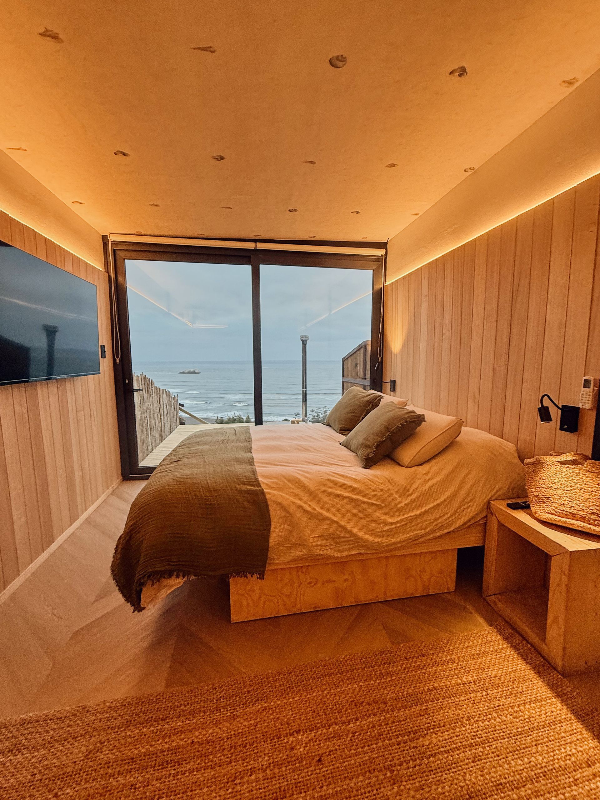 Modern bedroom with warm wood walls, large window, and ocean view at sunset.