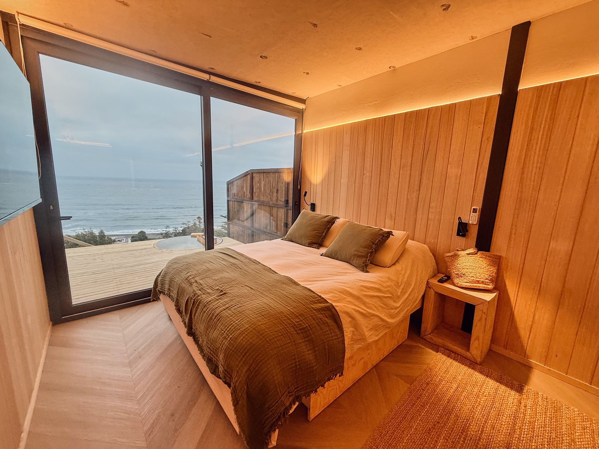 Modern bedroom with a large bed, warm wood interior, and floor-to-ceiling window overlooking the ocean