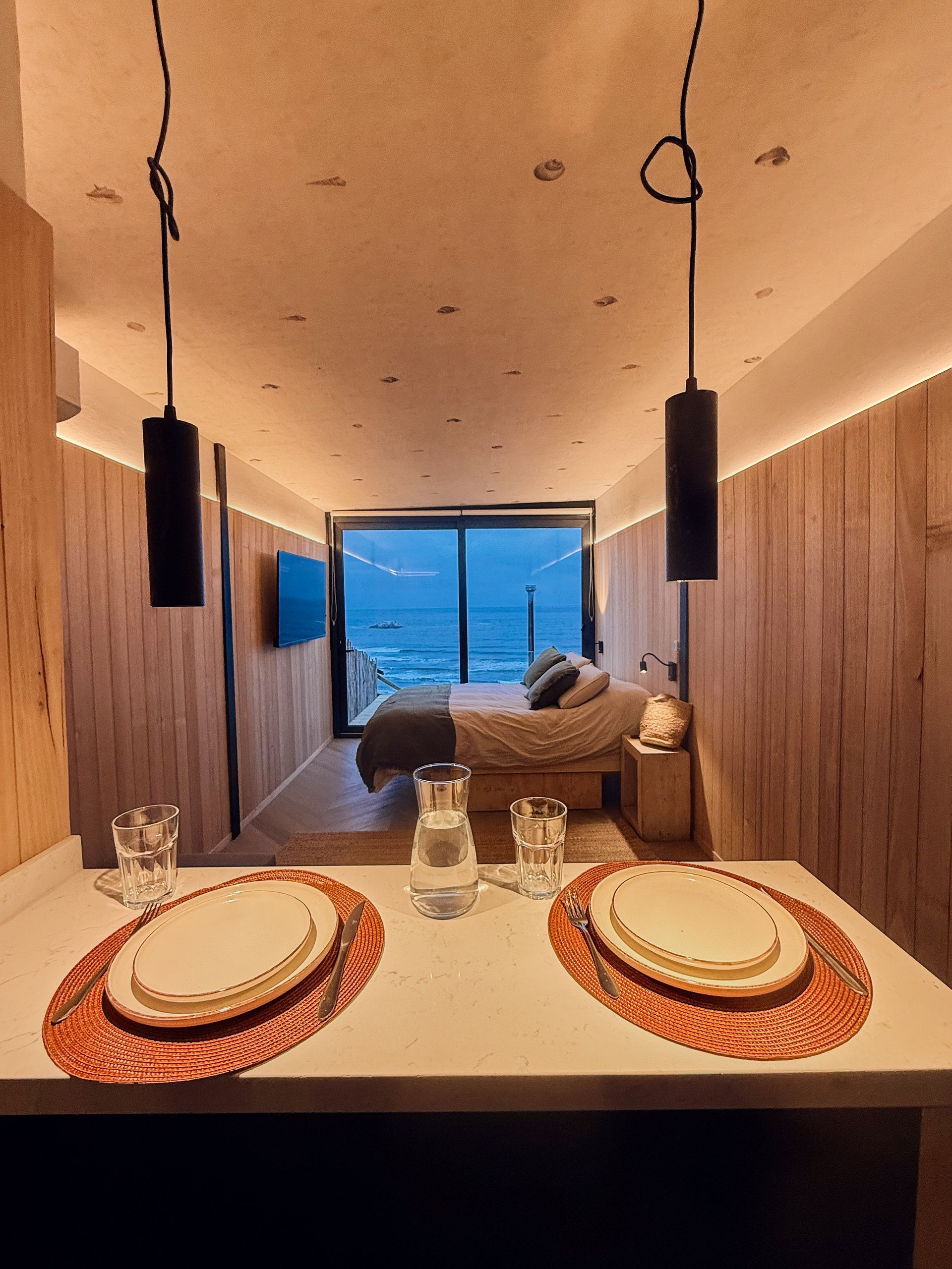 Modern dining area with wooden walls, a set table, pendant lights, and a blue-lit seating nook beyond.