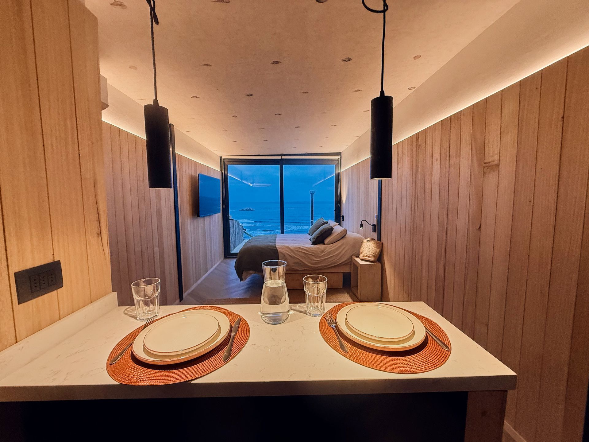 Modern dining nook with set table, wood-paneled walls, hanging lights, and a blue window view