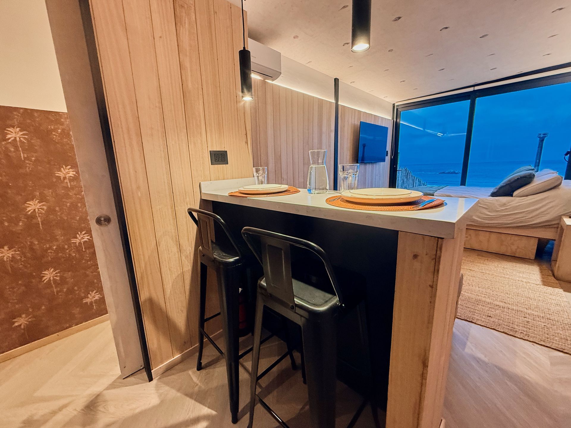 Modern kitchen island with two stools, pendant lights, and a bedroom by a large blue-tinted window.