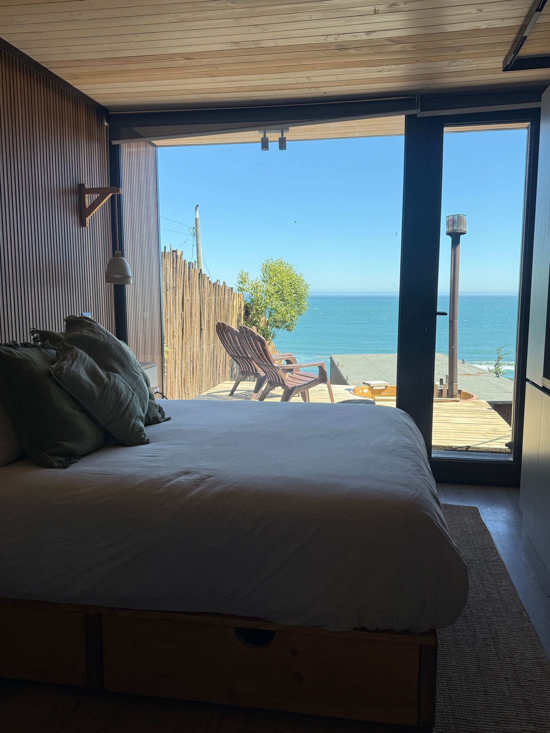 Cozy bedroom with a bed overlooking a patio, lounge chairs, and the ocean beyond a glass door