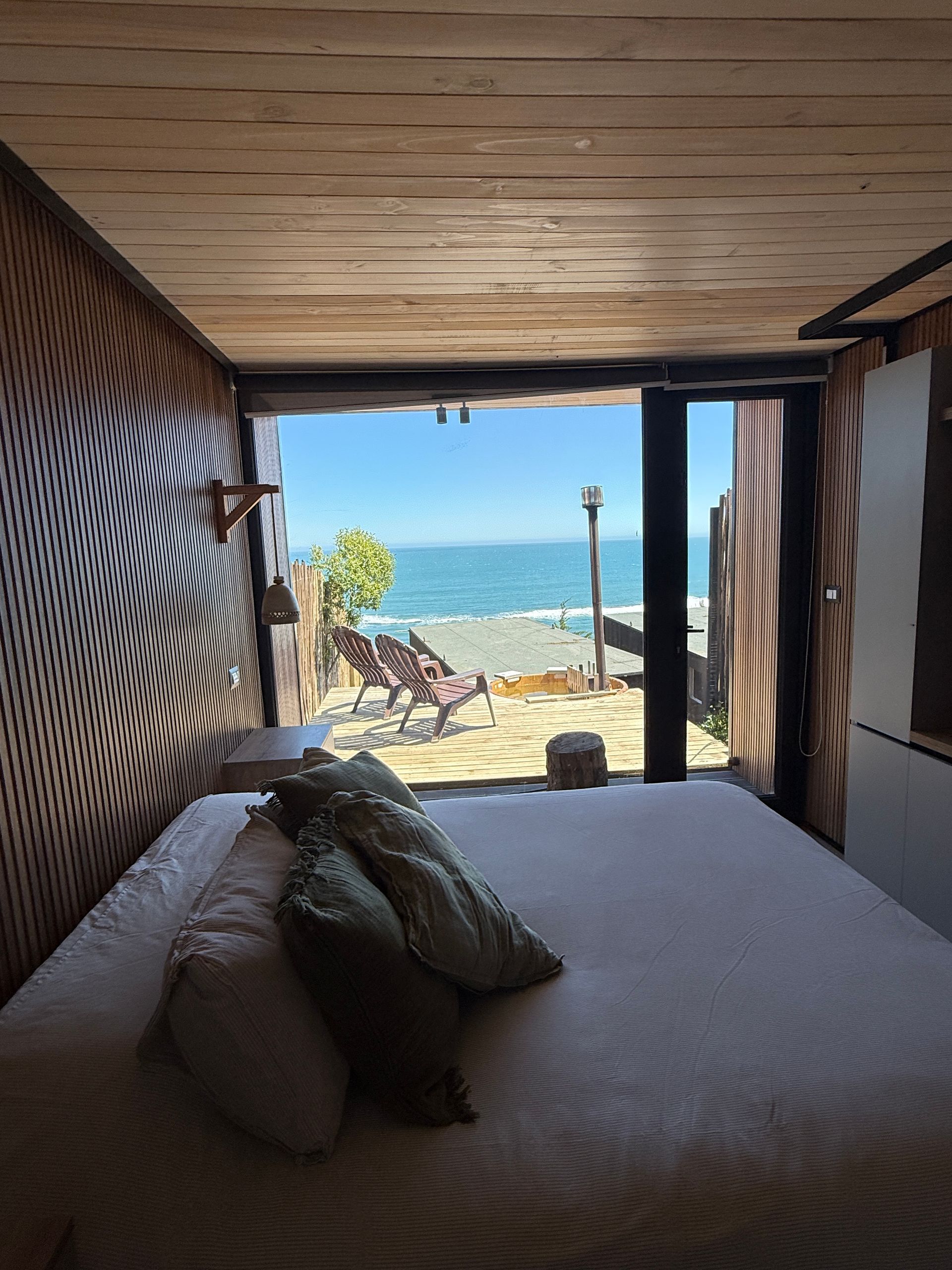 Cozy bedroom opening to a sunny beach and ocean view, with lounge chairs on the deck.