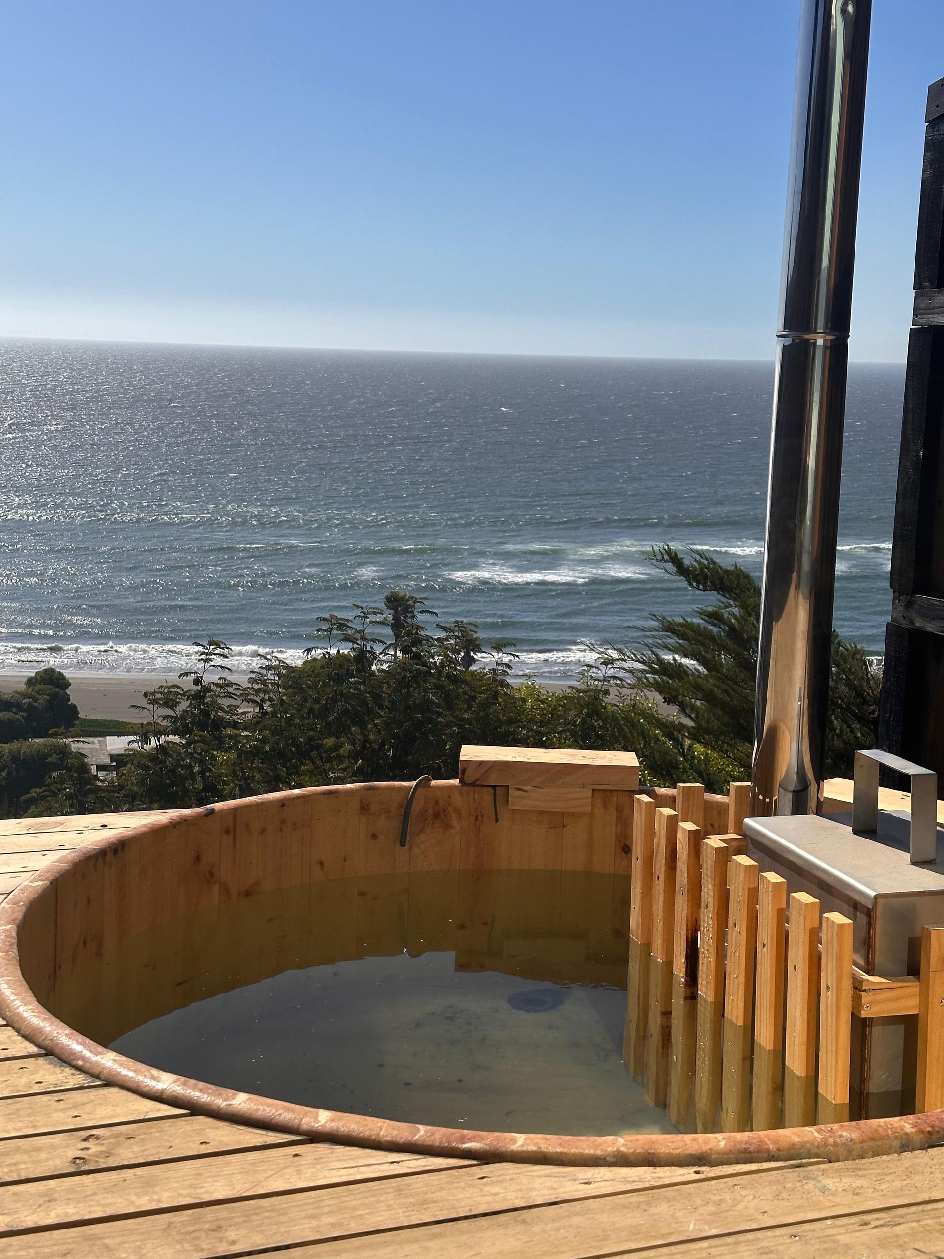 Wooden hot tub overlooking the ocean, with a sunny beach and blue sky in the background