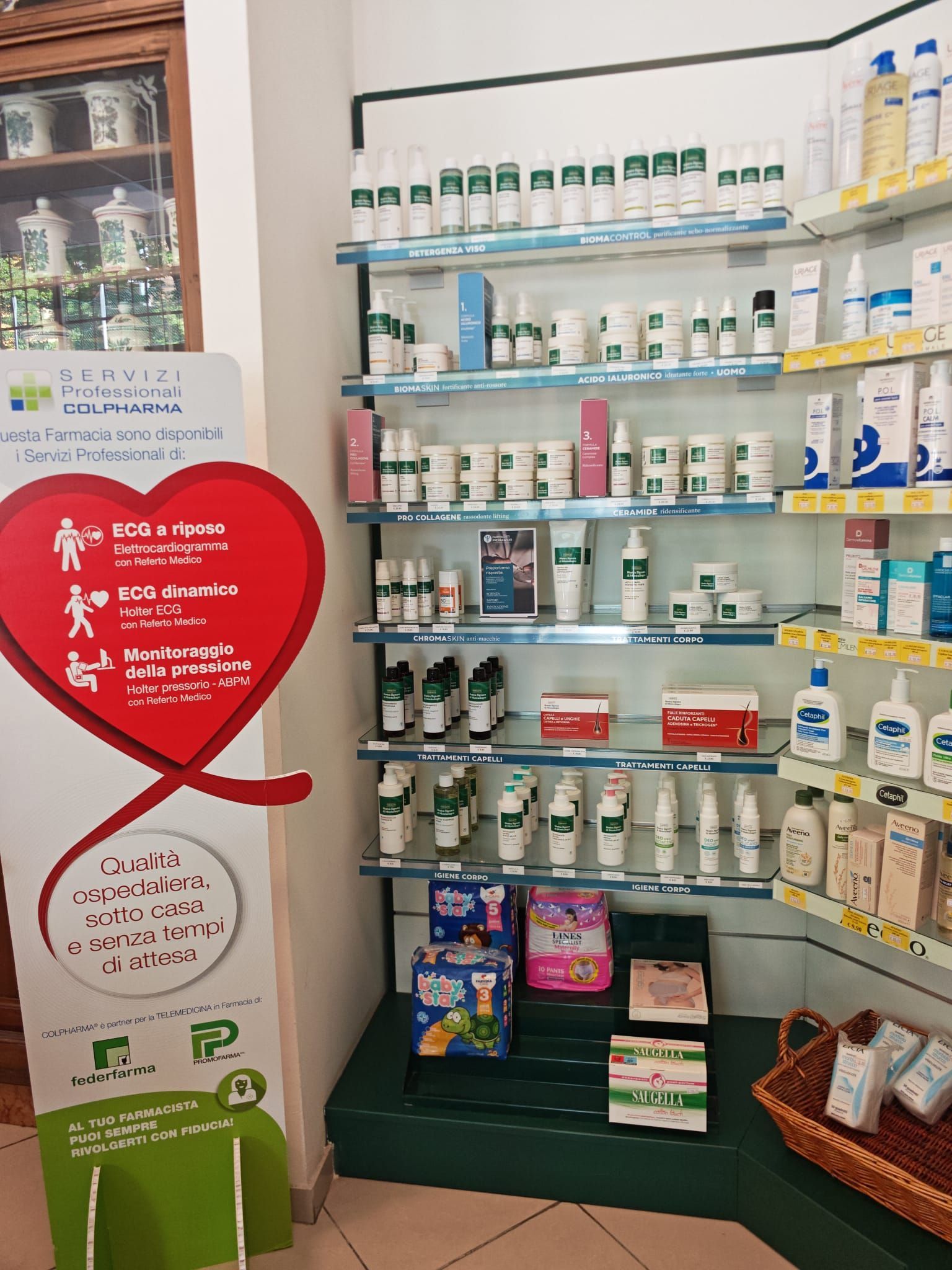 Scaffale della farmacia con medicinali e integratori, un cartello rosso a forma di cuore e espositori di prodotti.