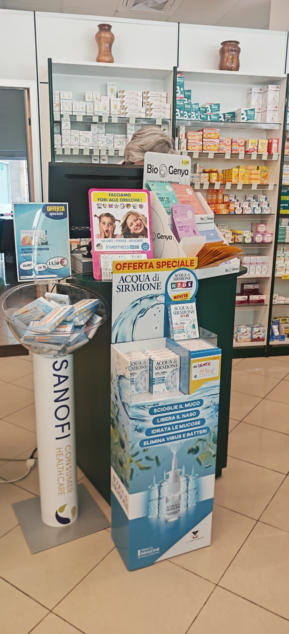 Esposizione in negozio di prodotti per la cura della pelle e la bellezza su uno scaffale e un piedistallo bianchi, con confezioni e brochure dai colori pastello.