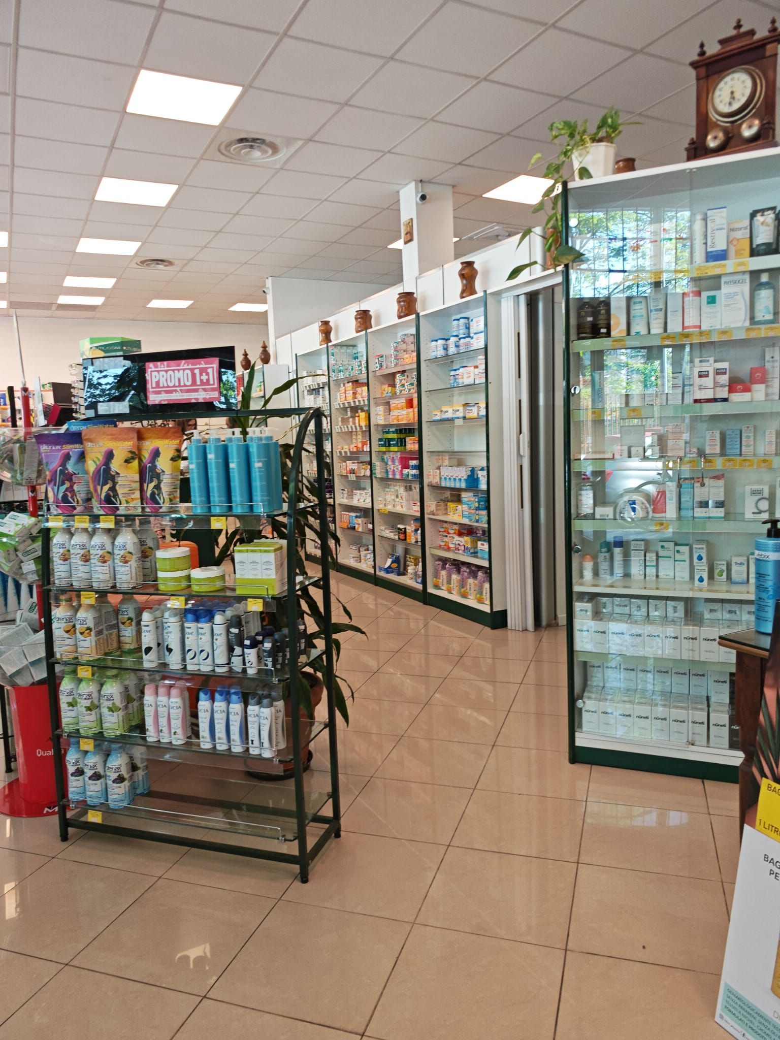Un corridoio di farmacia con scaffali pieni di articoli da toeletta, espositori di medicinali e una vetrina sotto luci intense del soffitto.