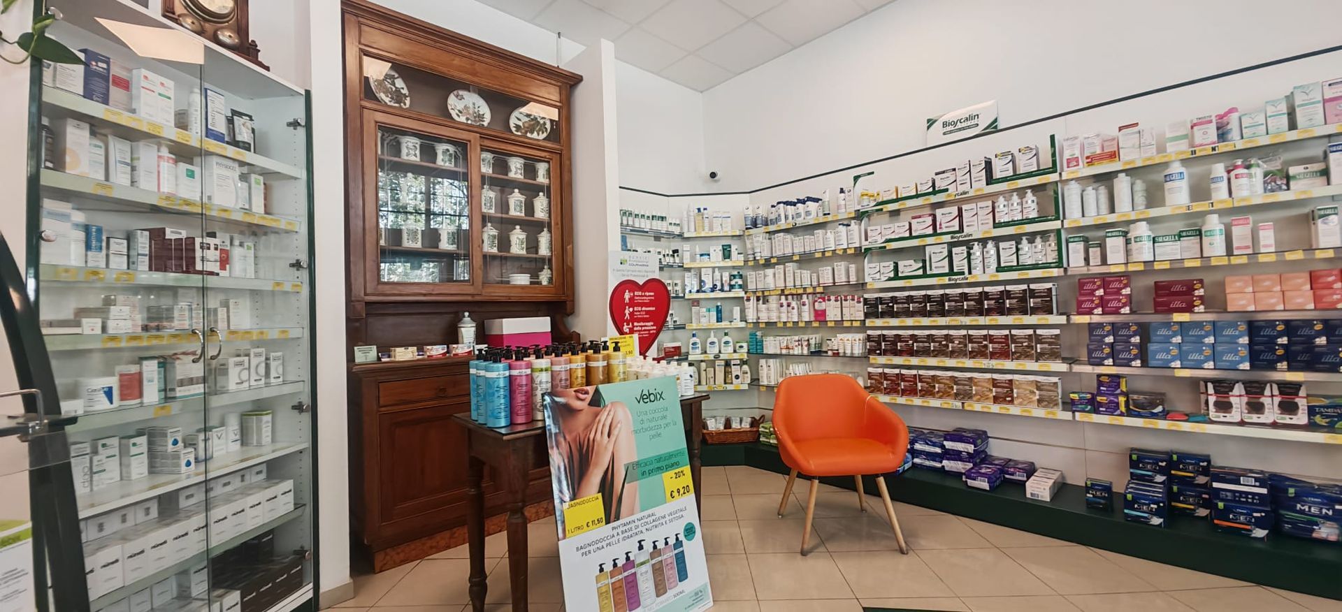 Interno luminoso di una farmacia con scaffali pieni di medicinali, una vetrina, una sedia arancione e un bancone espositivo.