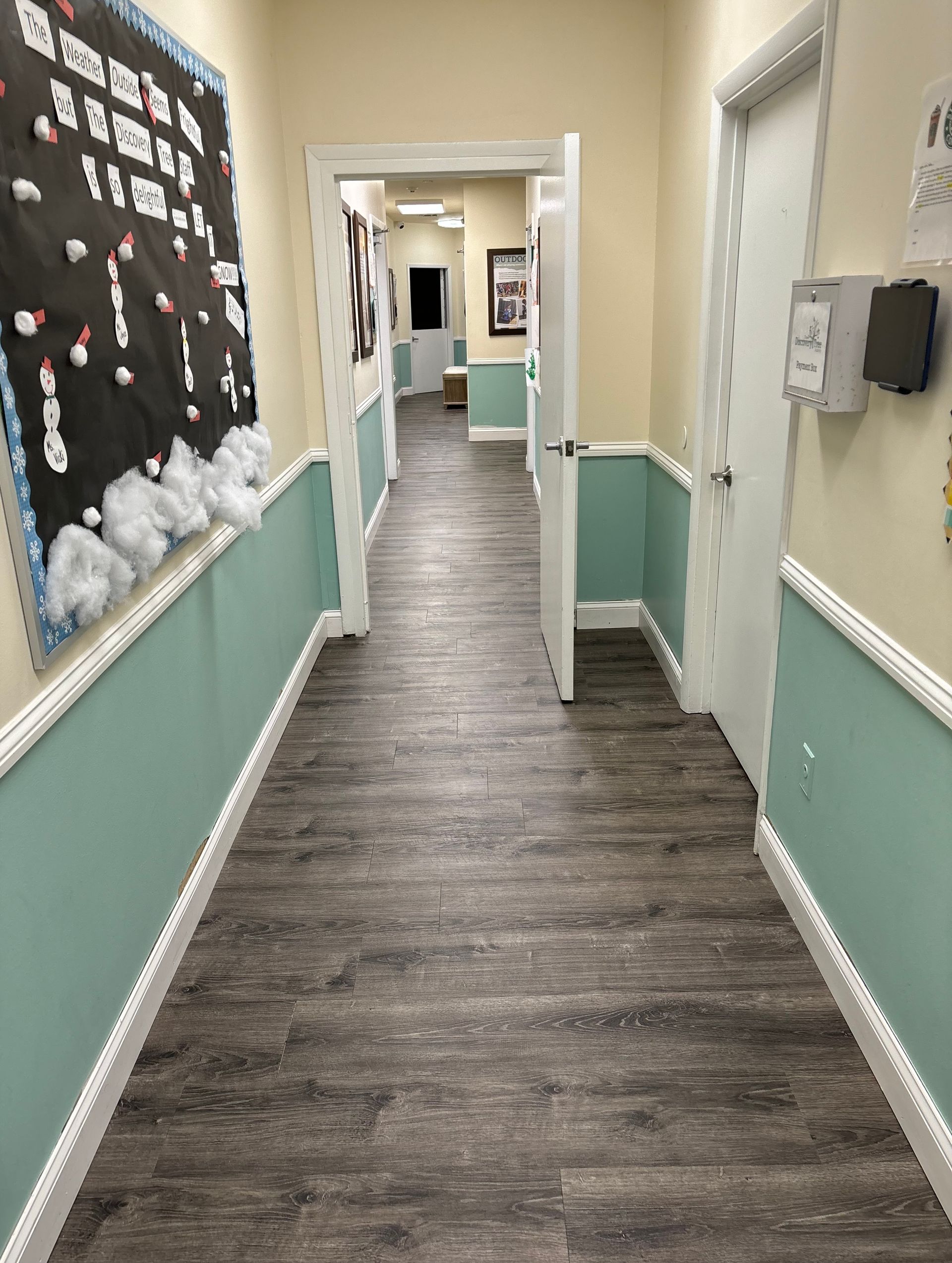 A hallway with wood-look flooring, light walls, and teal wainscoting leading to open doorways and a bulletin board.