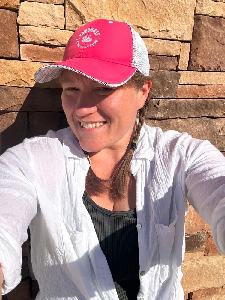 Woman in a white shirt and pink cap smiles in front of a stone wall.