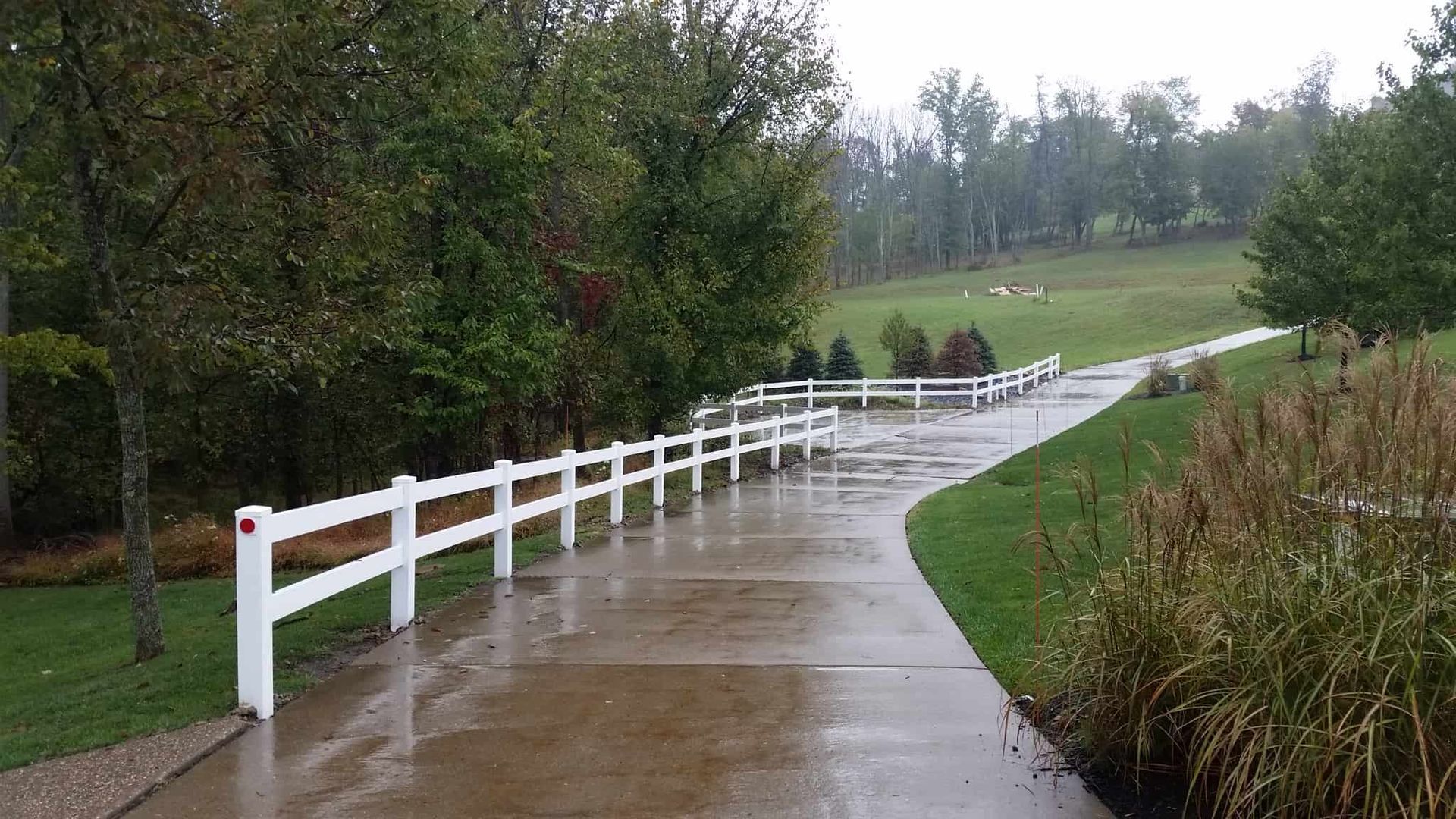 Rainy paved path with white fence curving through a green park
