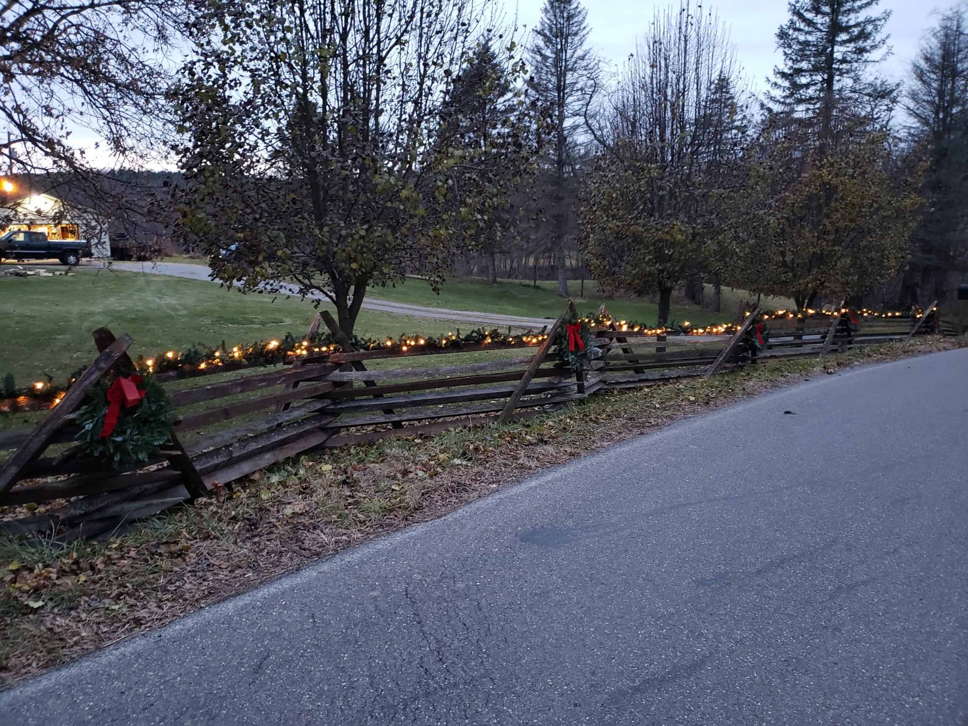 Holiday-lit wooden fence along a roadside at dusk, with trees and a house in the background