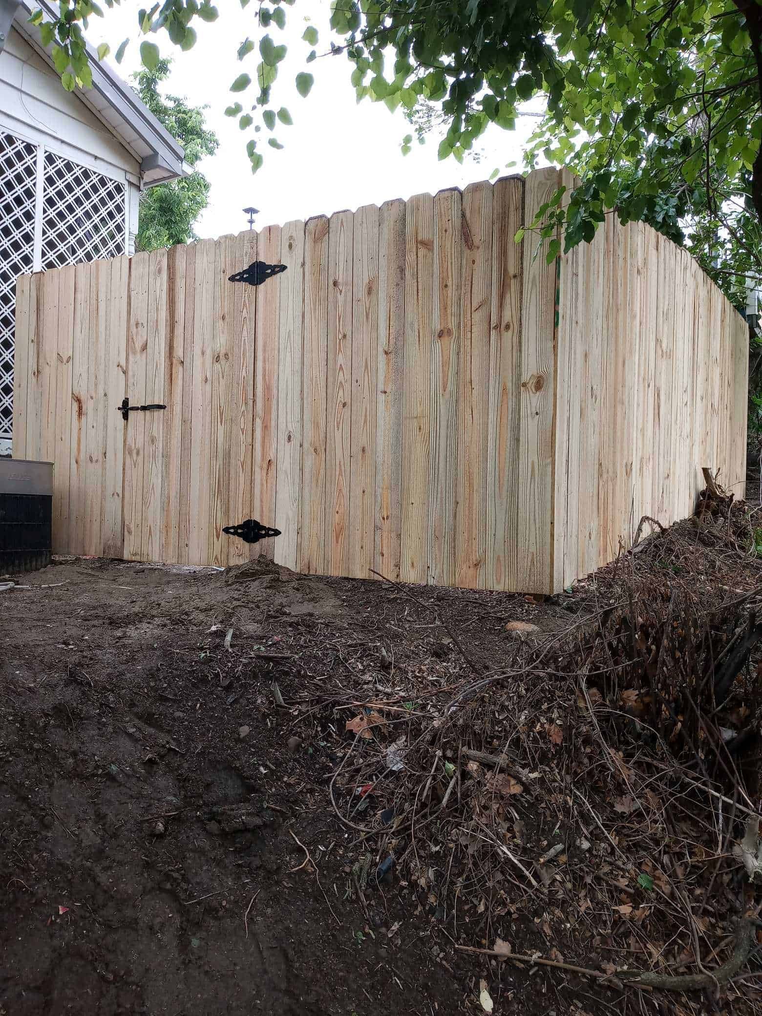 Wooden gate in a fenced yard beside a house, with dark mulch and leafy tree branches overhead.