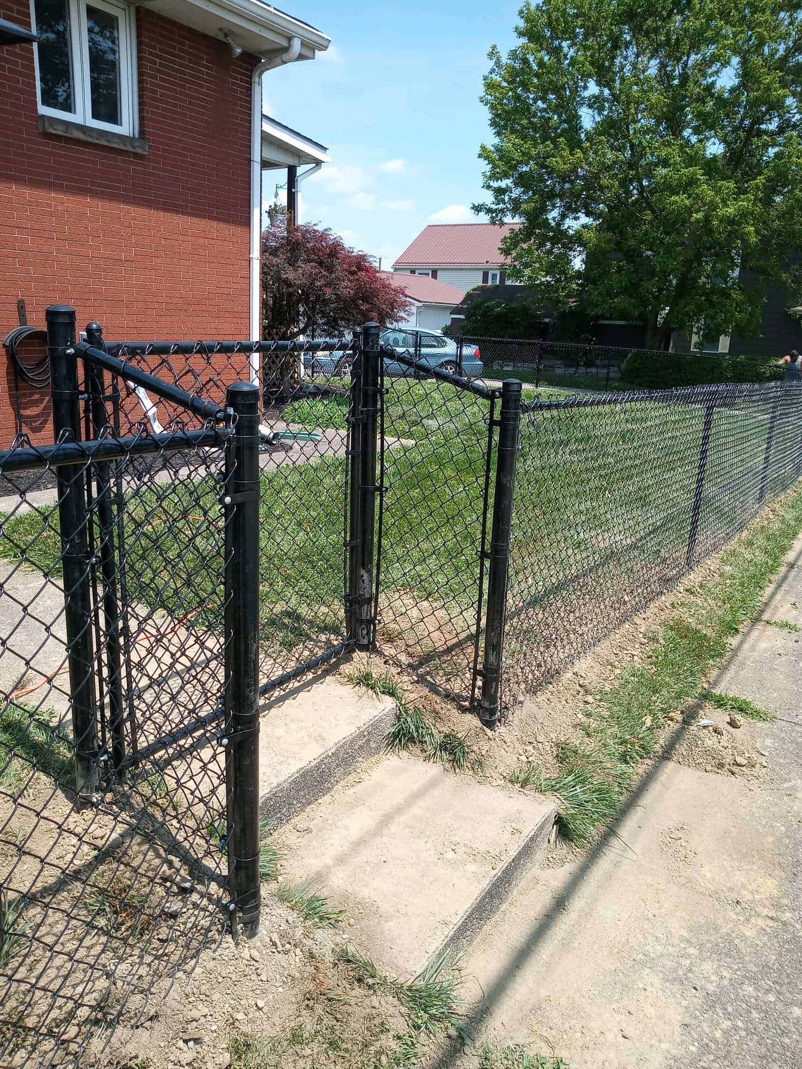 Black metal fence beside a red brick house along a sidewalk, with grass and trees in the yard.