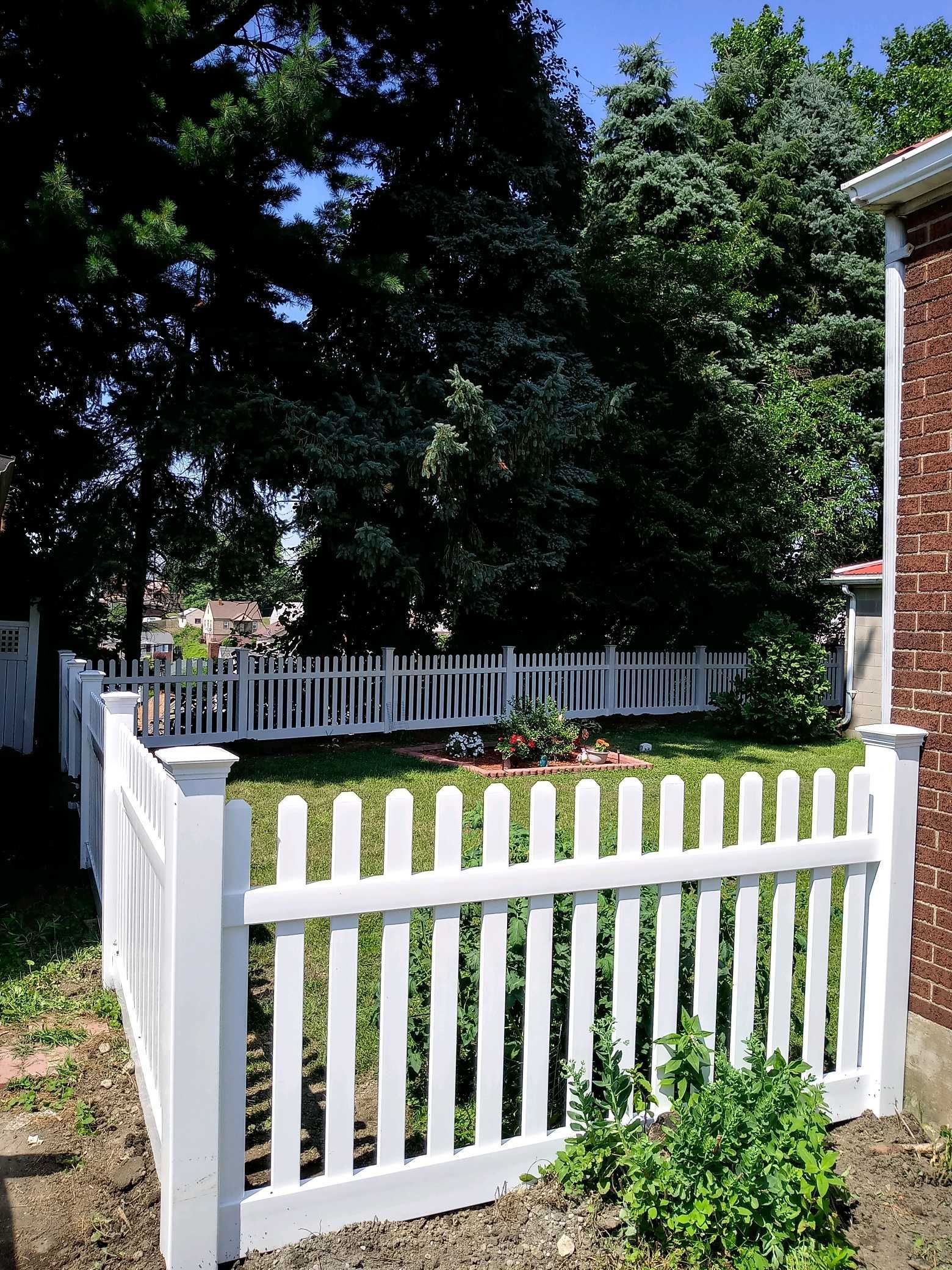 White picket fence around a small front yard with green grass, trees, and a brick house in the background