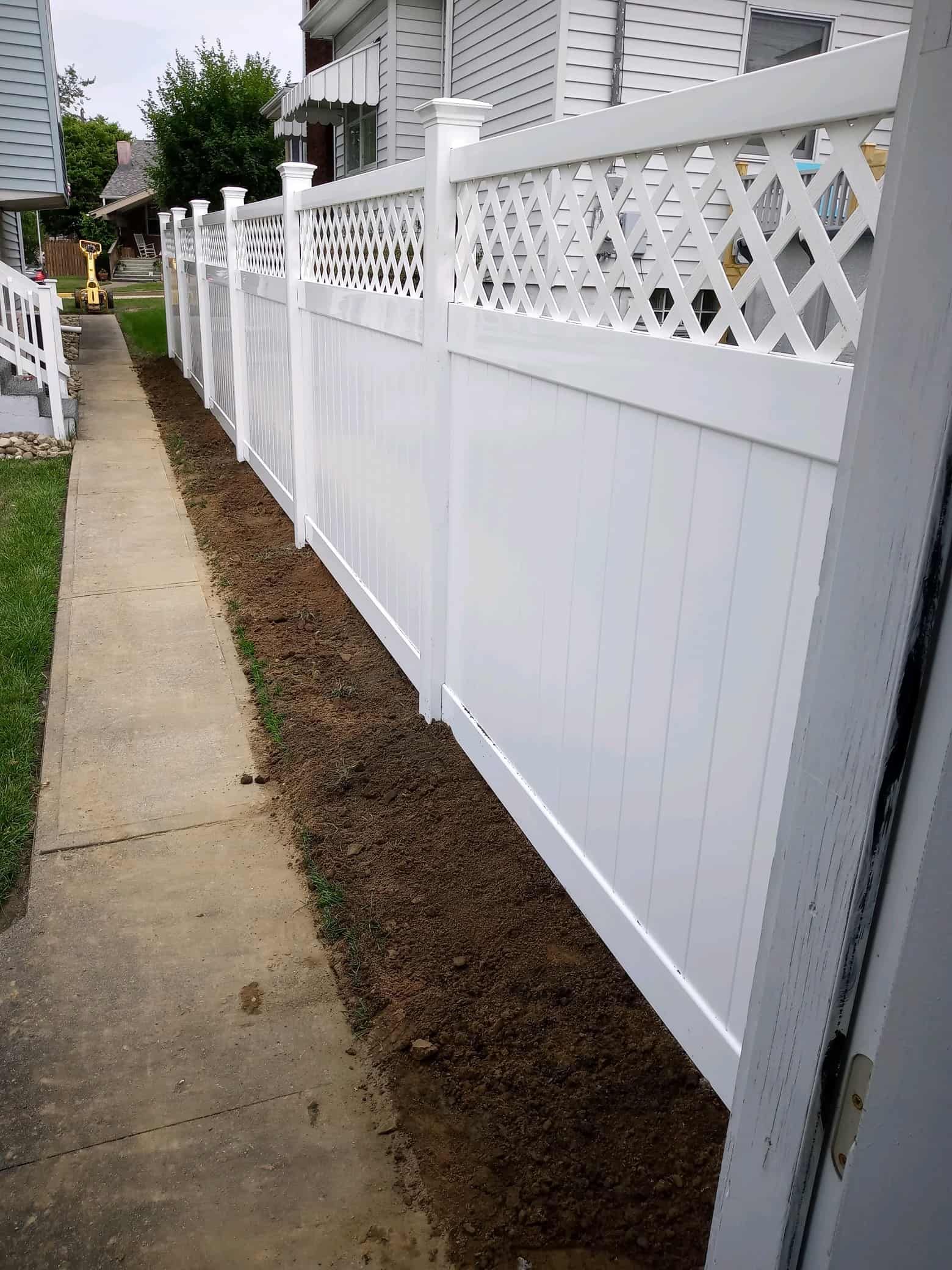 Narrow sidewalk beside a white vinyl fence with lattice top and a small dirt strip along the base