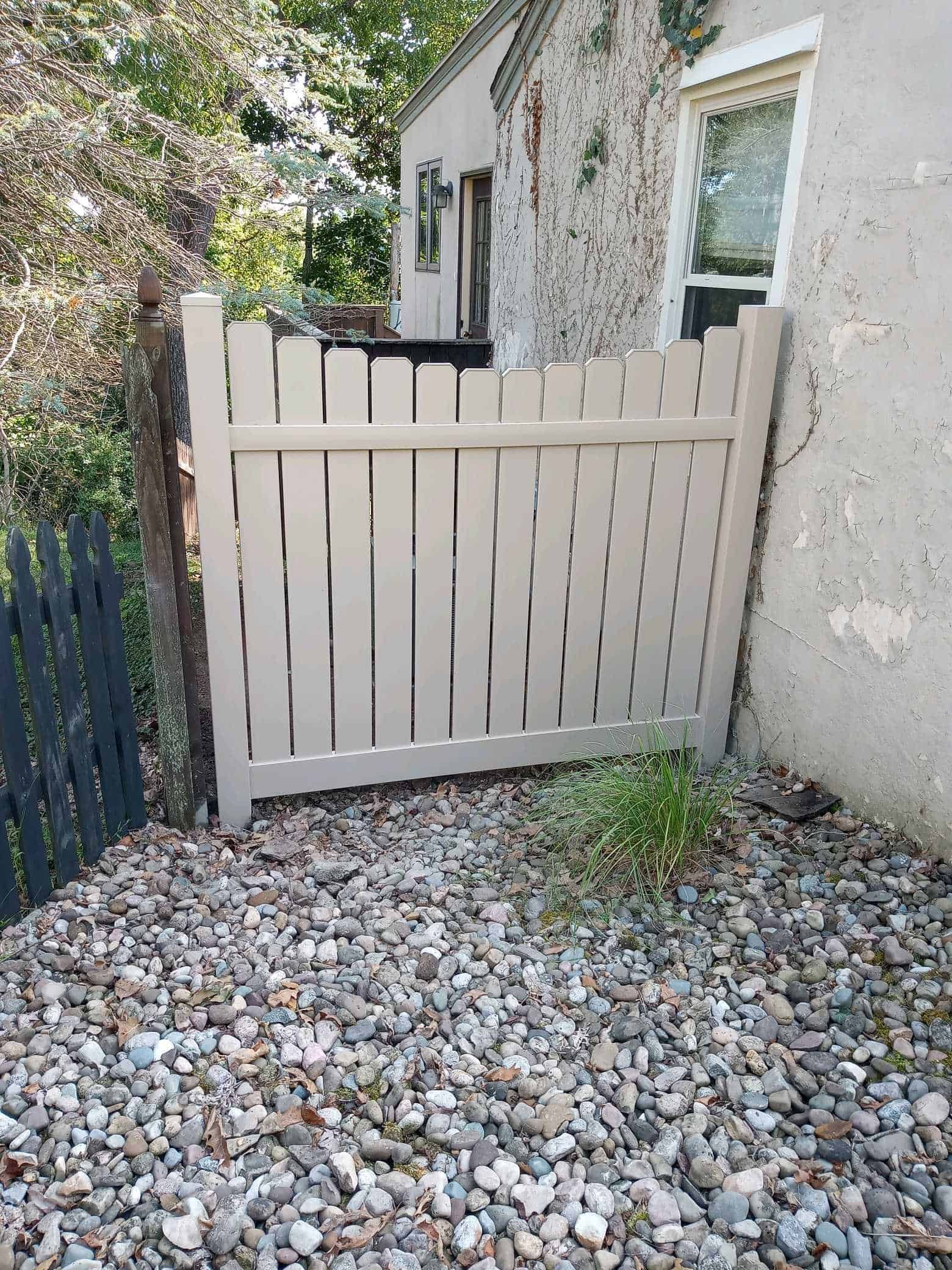 Beige wooden gate beside a stone house, with gravel ground and greenery