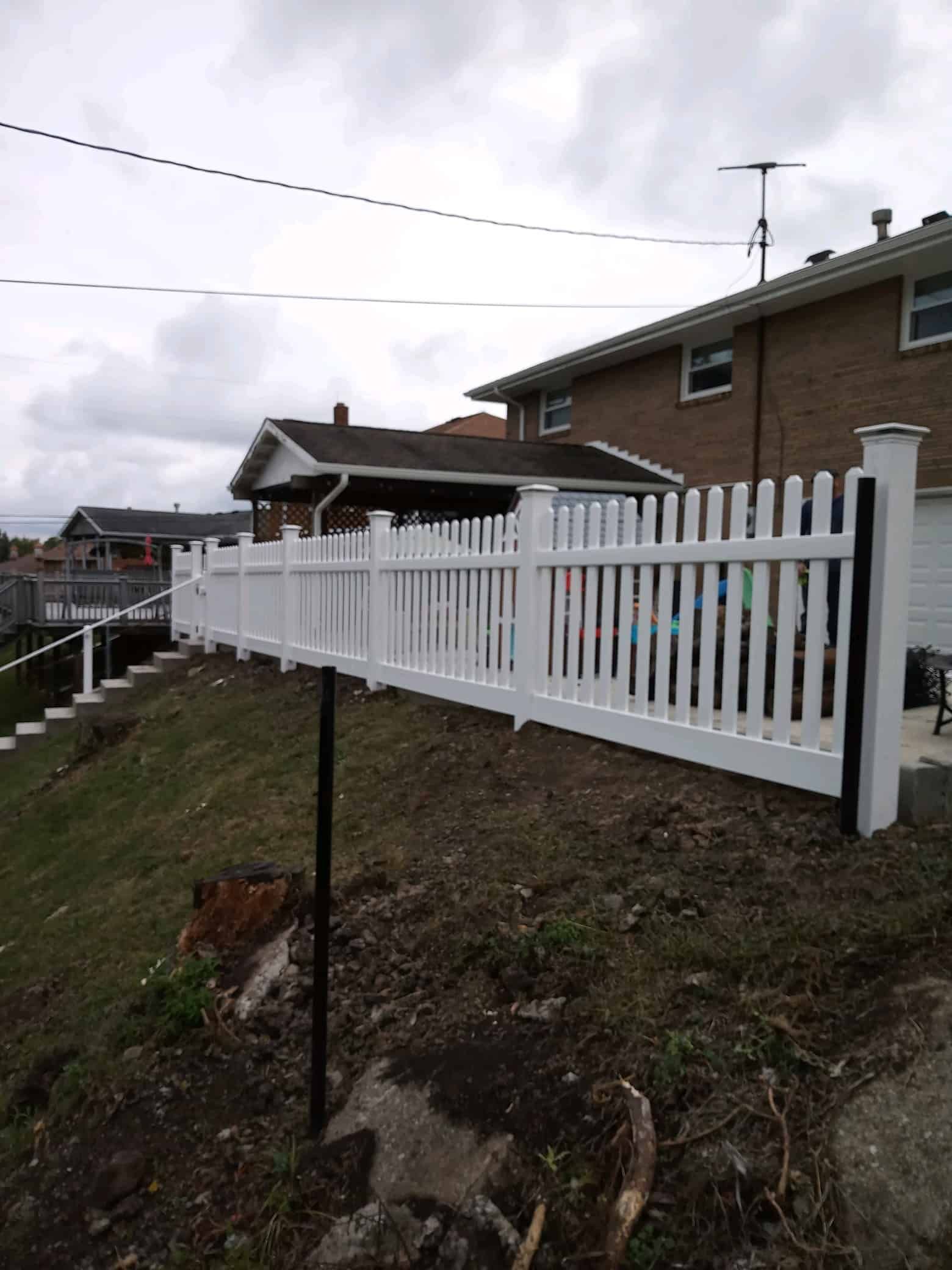 White fence on a sloped yard beside a brick house under a cloudy sky.