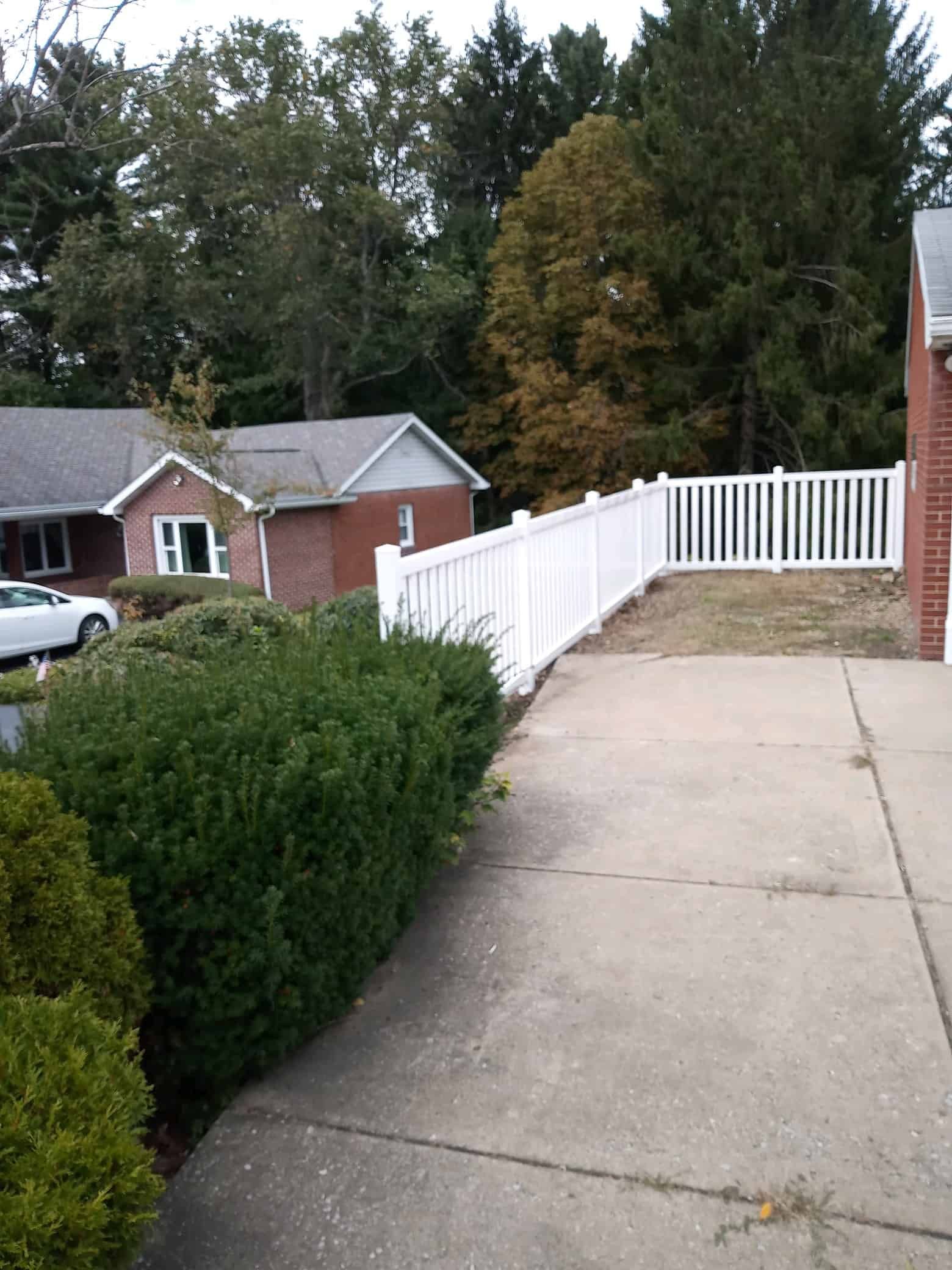 Concrete driveway beside hedge and white fence leading to a brick house in a wooded neighborhood