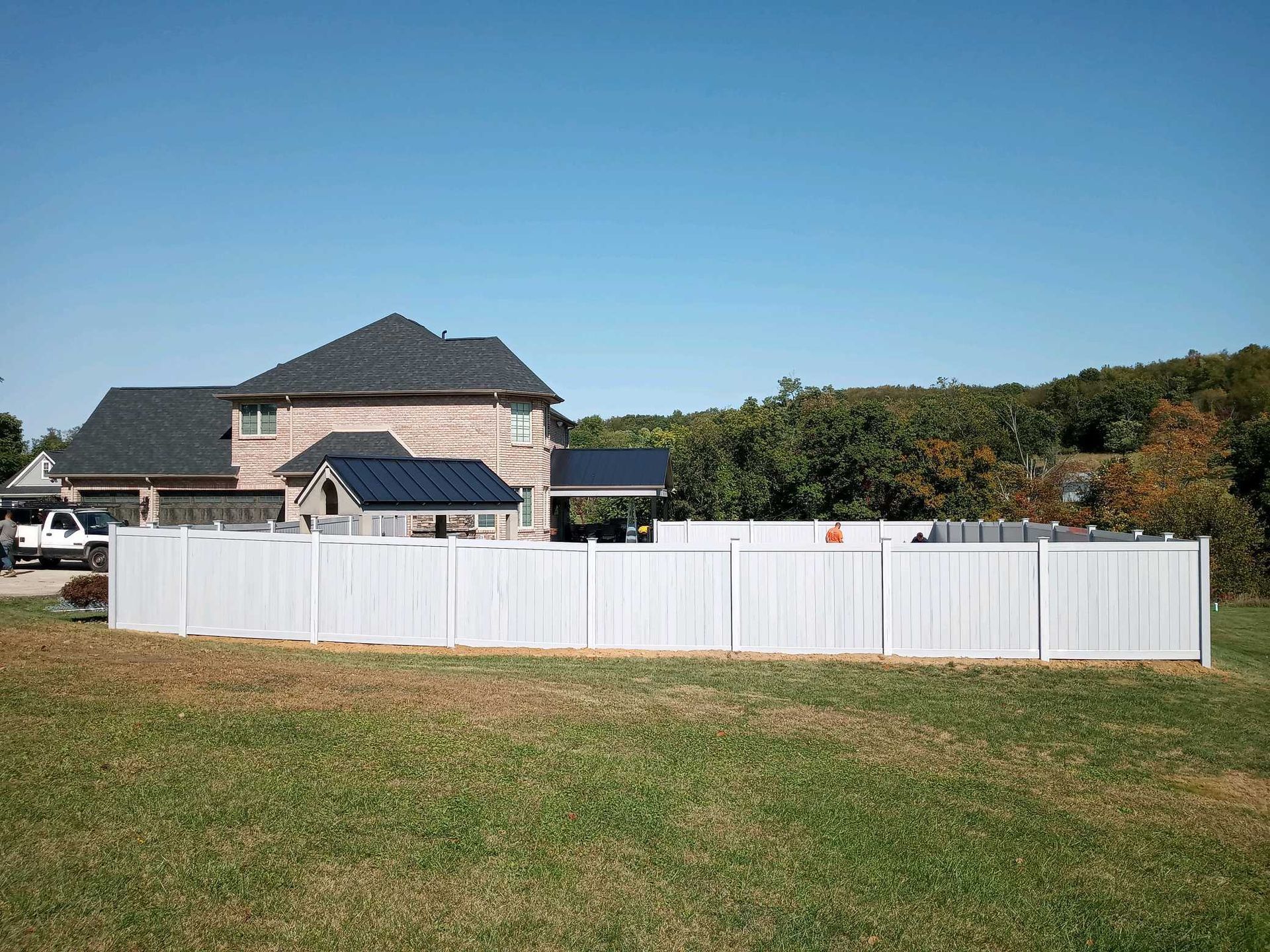 White fence in front of a large house with a dark roof, grassy foreground, and blue sky.