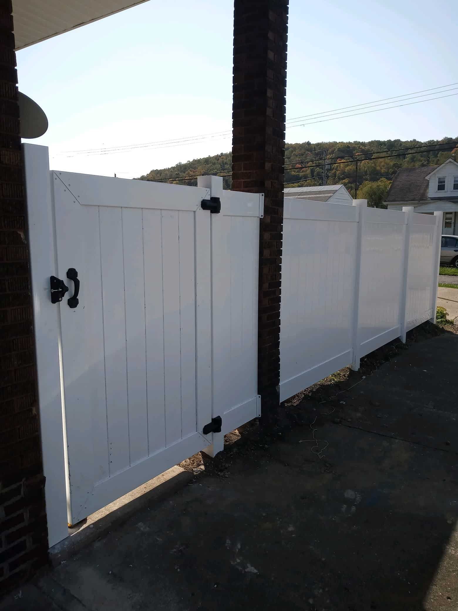 White wooden gate in a fenced driveway beside a brick post, with hills in the background.
