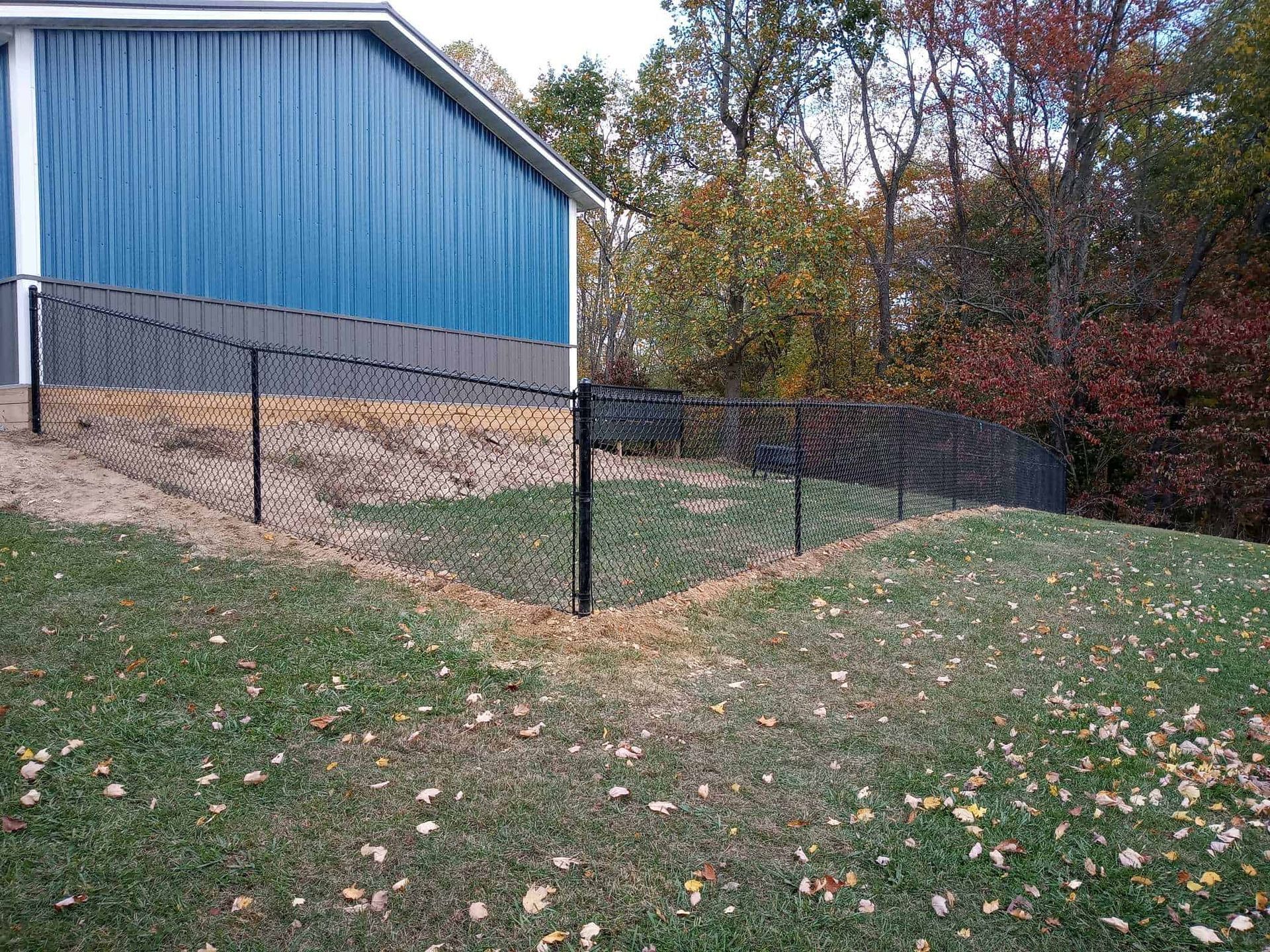 Blue building beside black chain-link fence on a grassy slope with autumn trees in the background