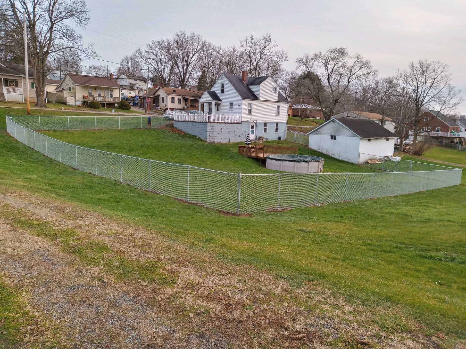 Chain-link-fenced grassy field beside white houses and a small outbuilding on a hill under bare trees.