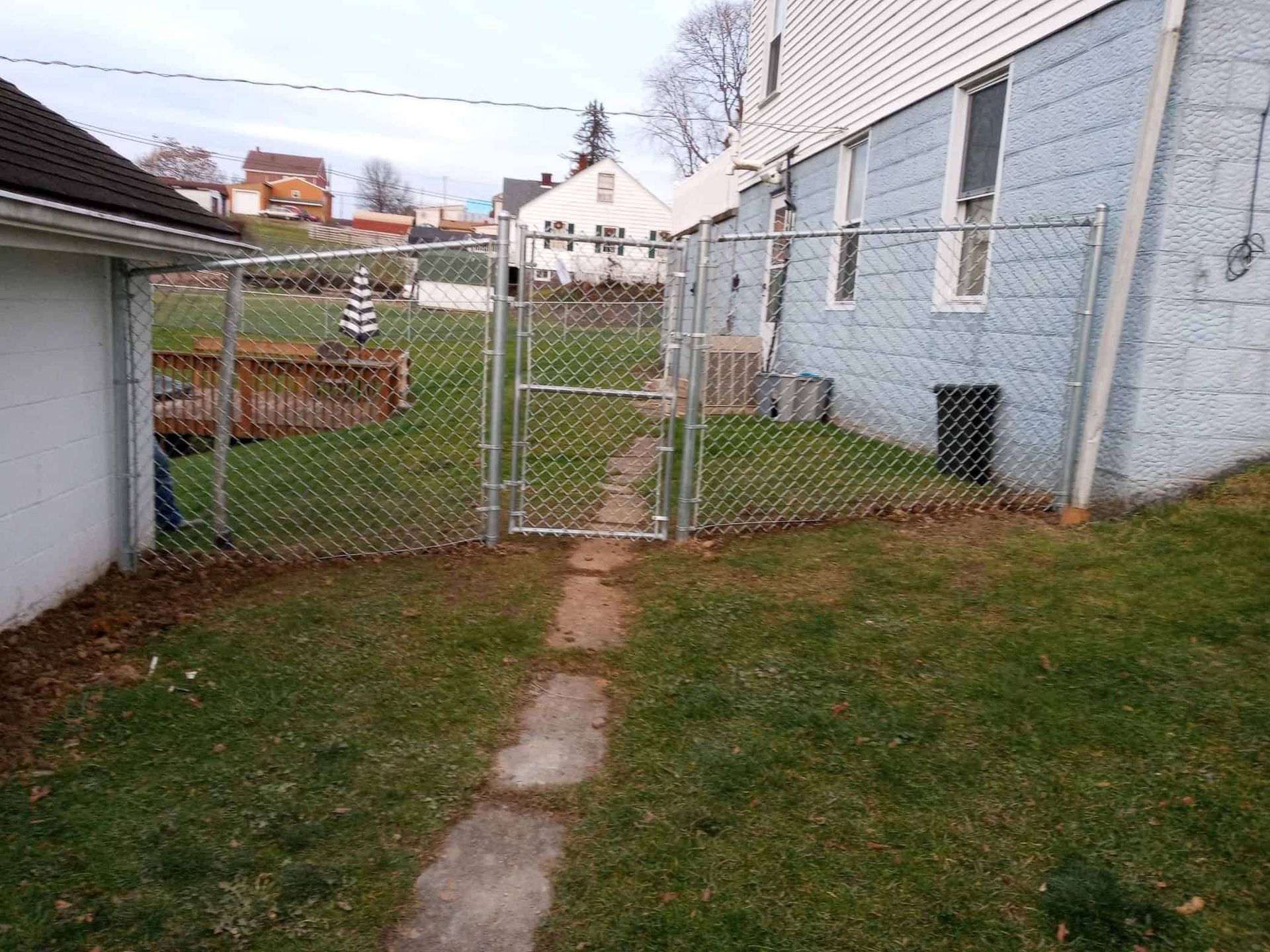 Narrow backyard path leading to a chain-link gate between two houses, with grass and a white building on the right