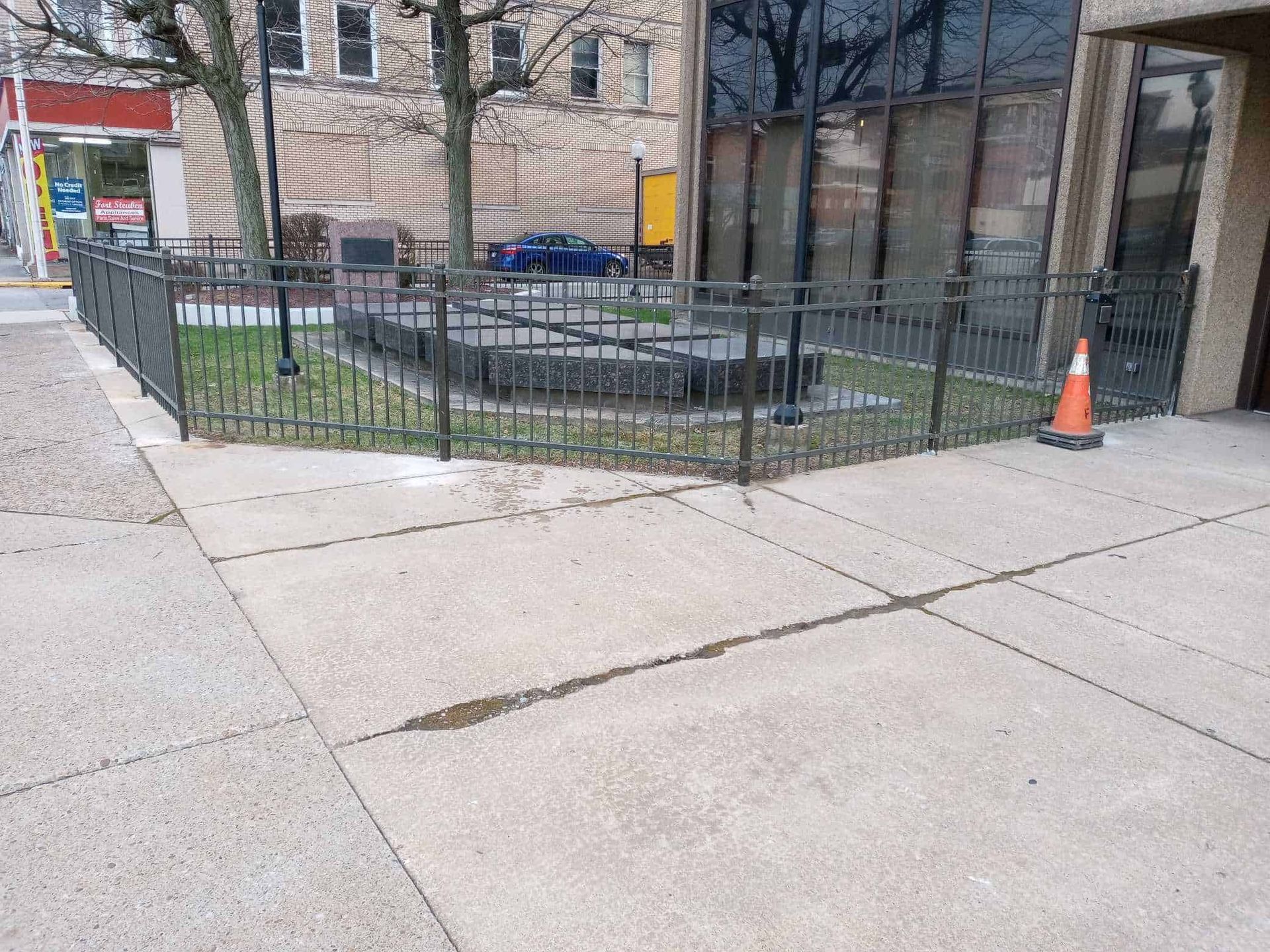 City sidewalk beside a fenced raised planter and buildings, with an orange traffic cone near the entrance.