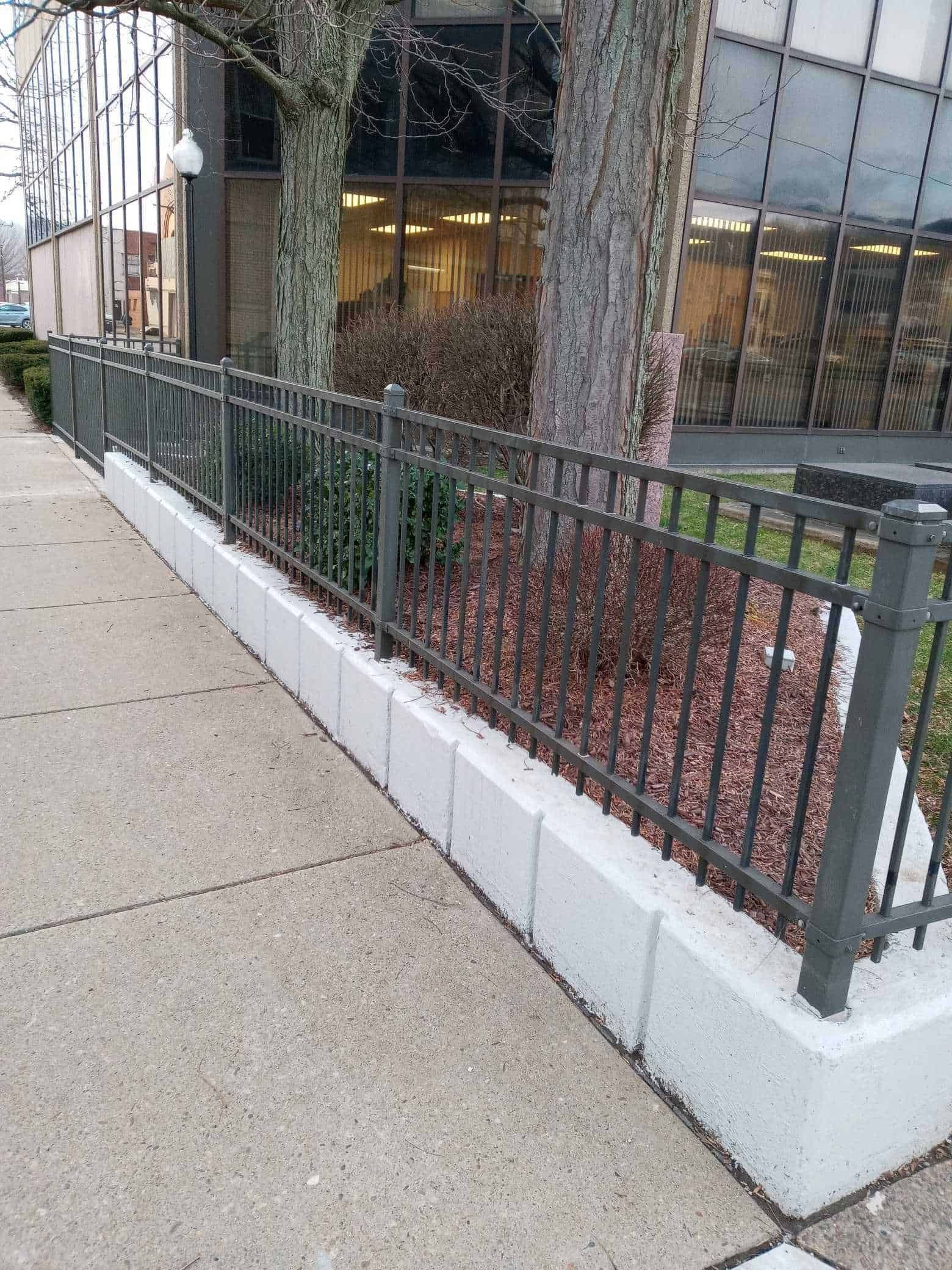 Sidewalk beside a building with a black metal fence and white concrete barrier around a landscaped bed