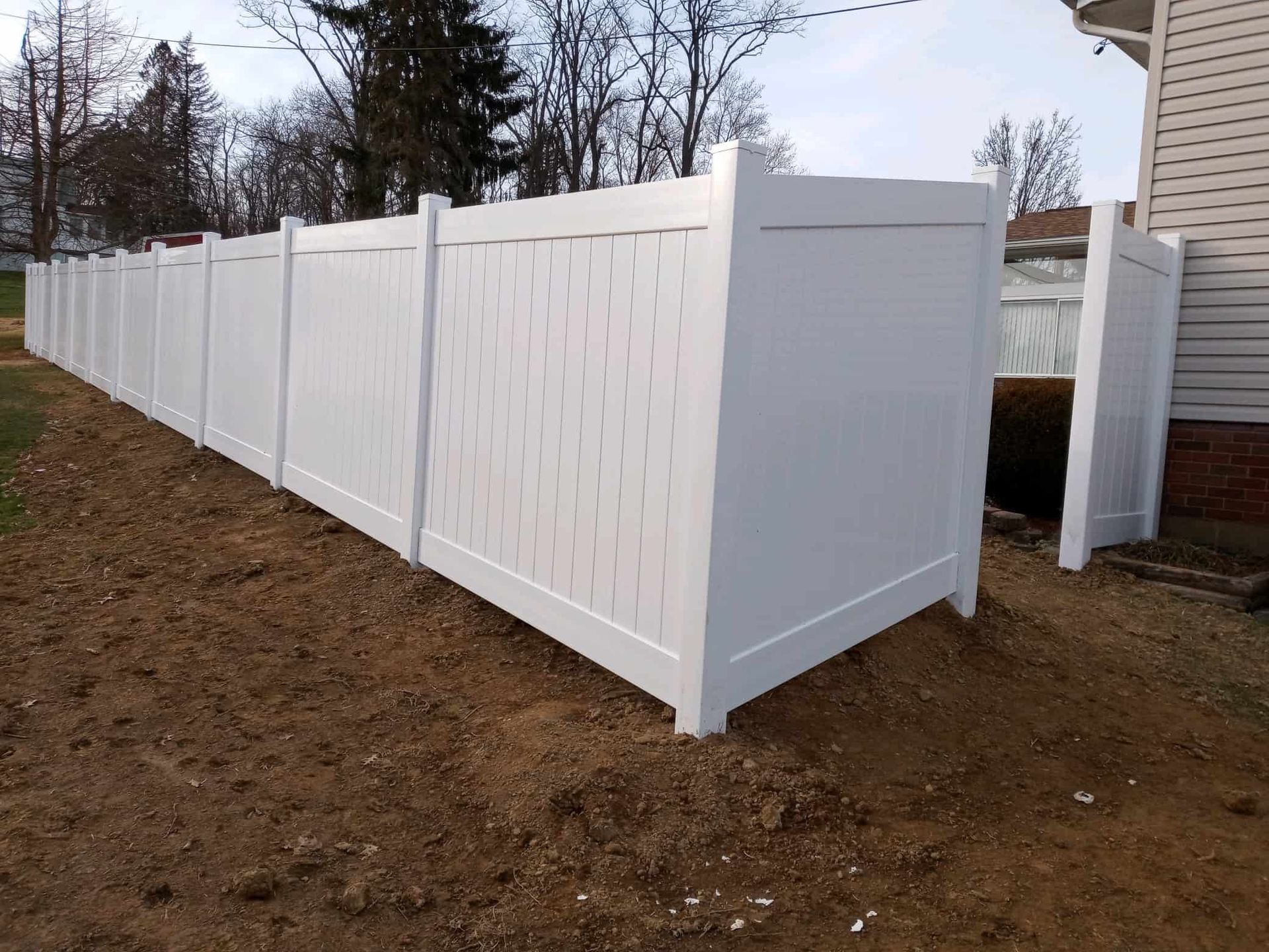White vinyl privacy fence running along a dirt yard beside a house and leafless trees