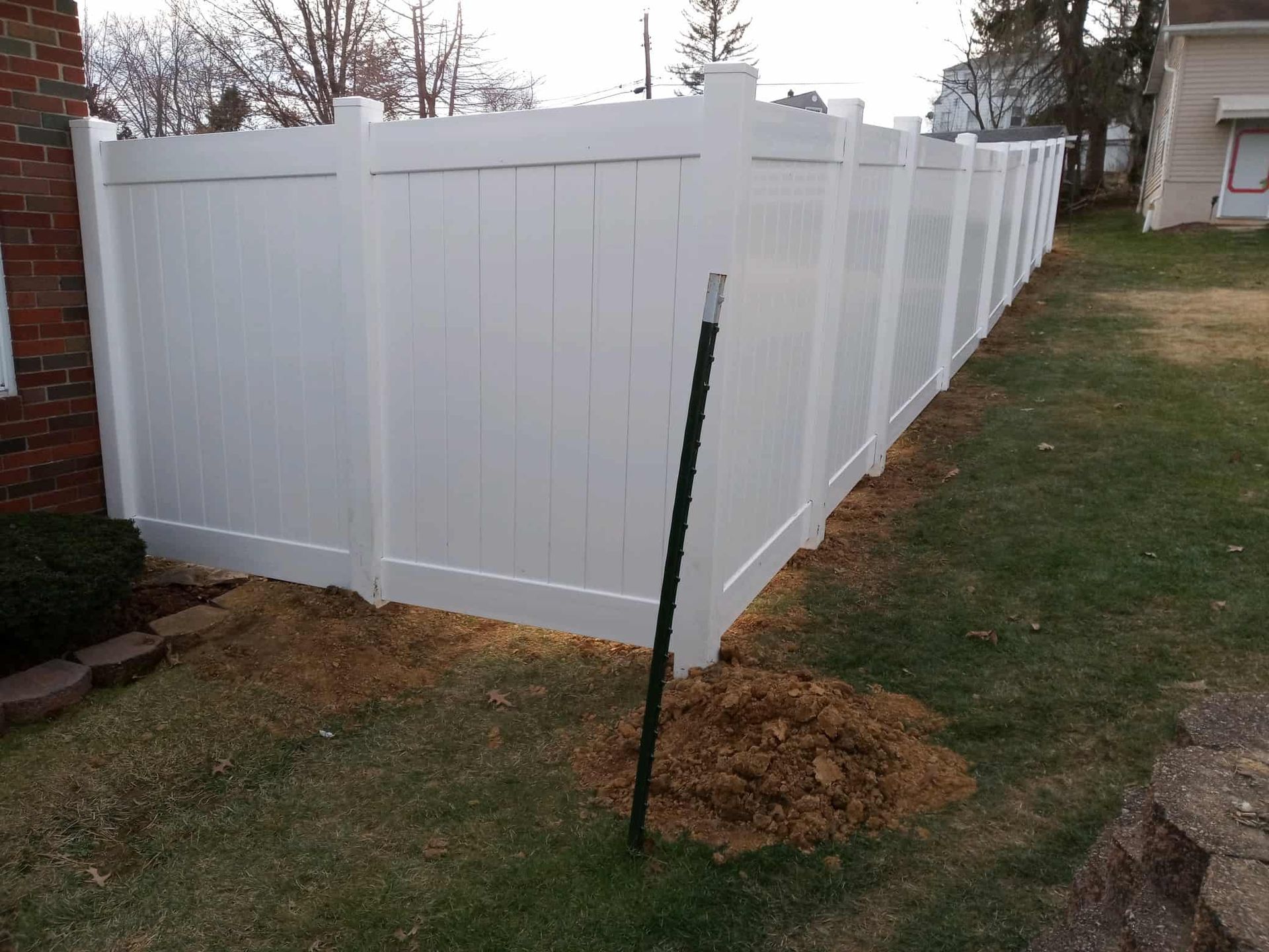 White vinyl fence along a grassy yard beside a brick house, with a young tree stake in front