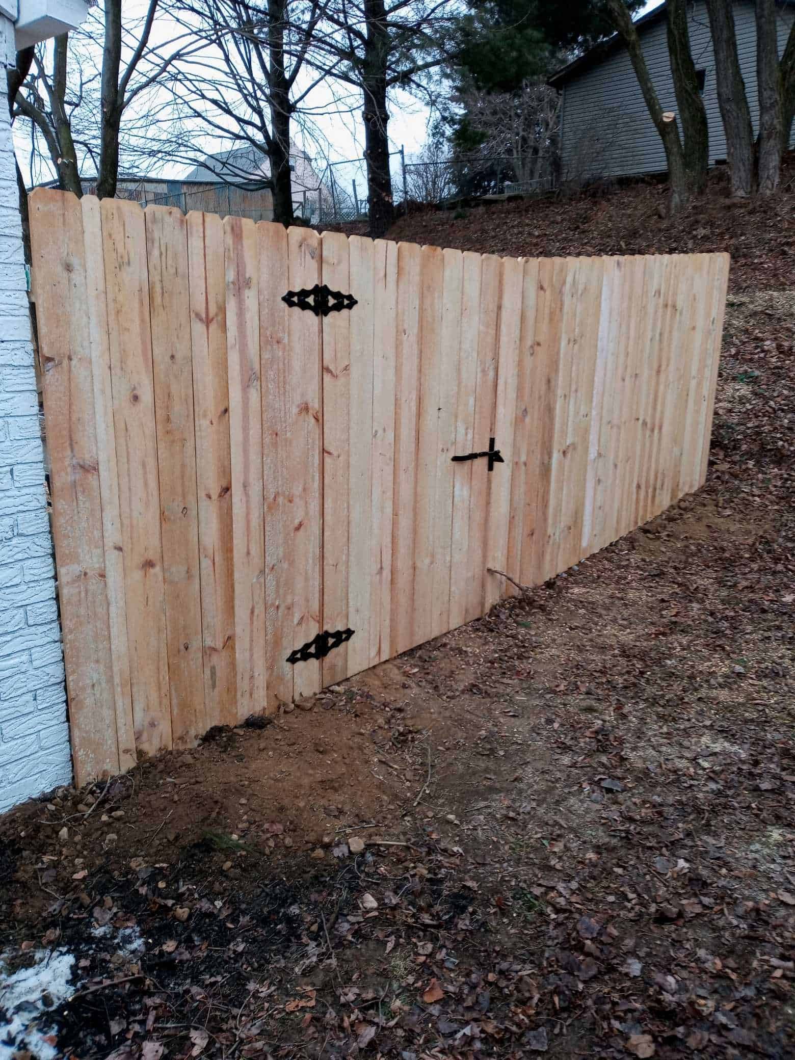 New wooden fence with black hinges beside a house on a wooded slope