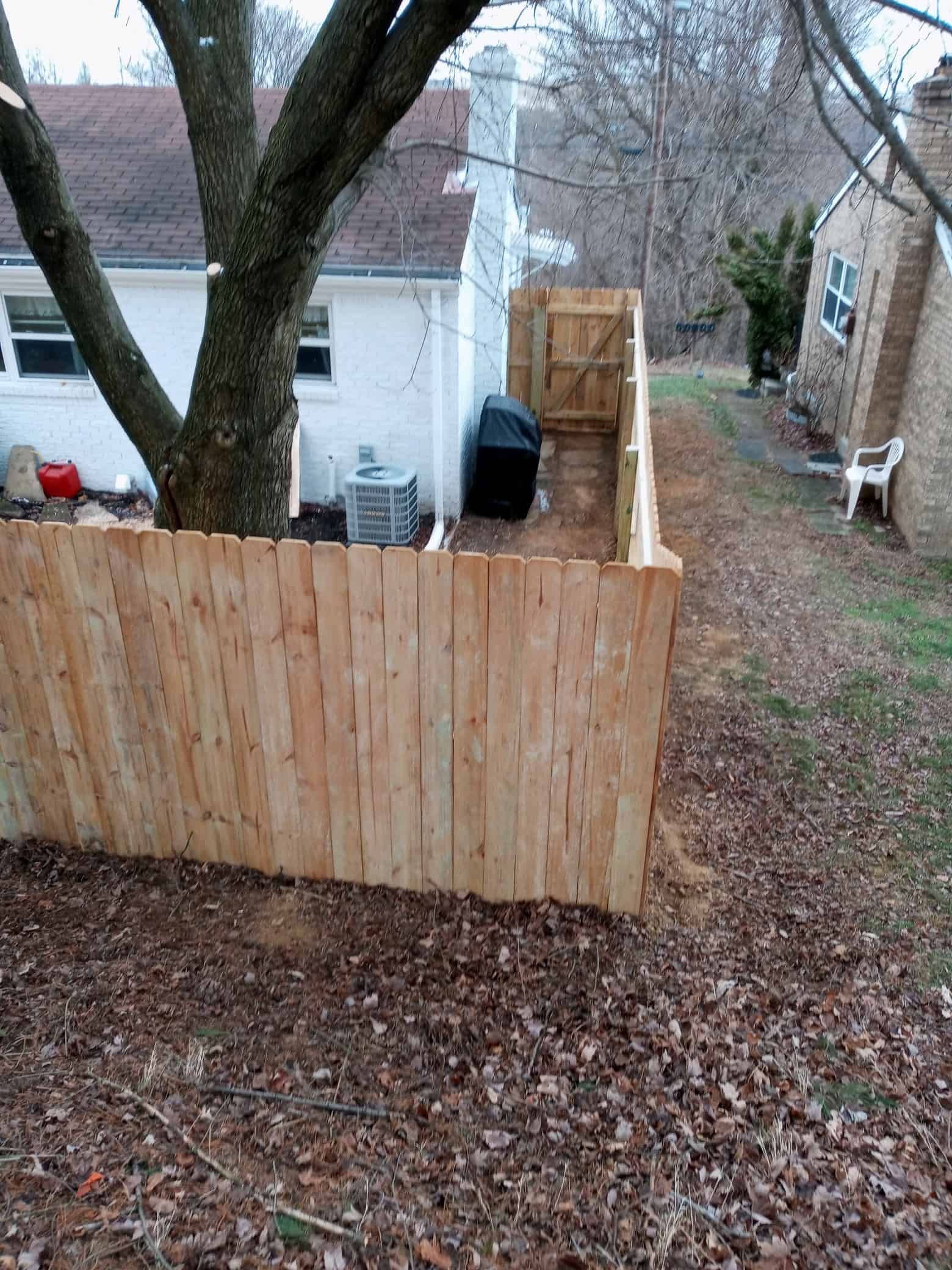 Wooden privacy fence around a backyard with trees, a white house, and a black grill on a deck.