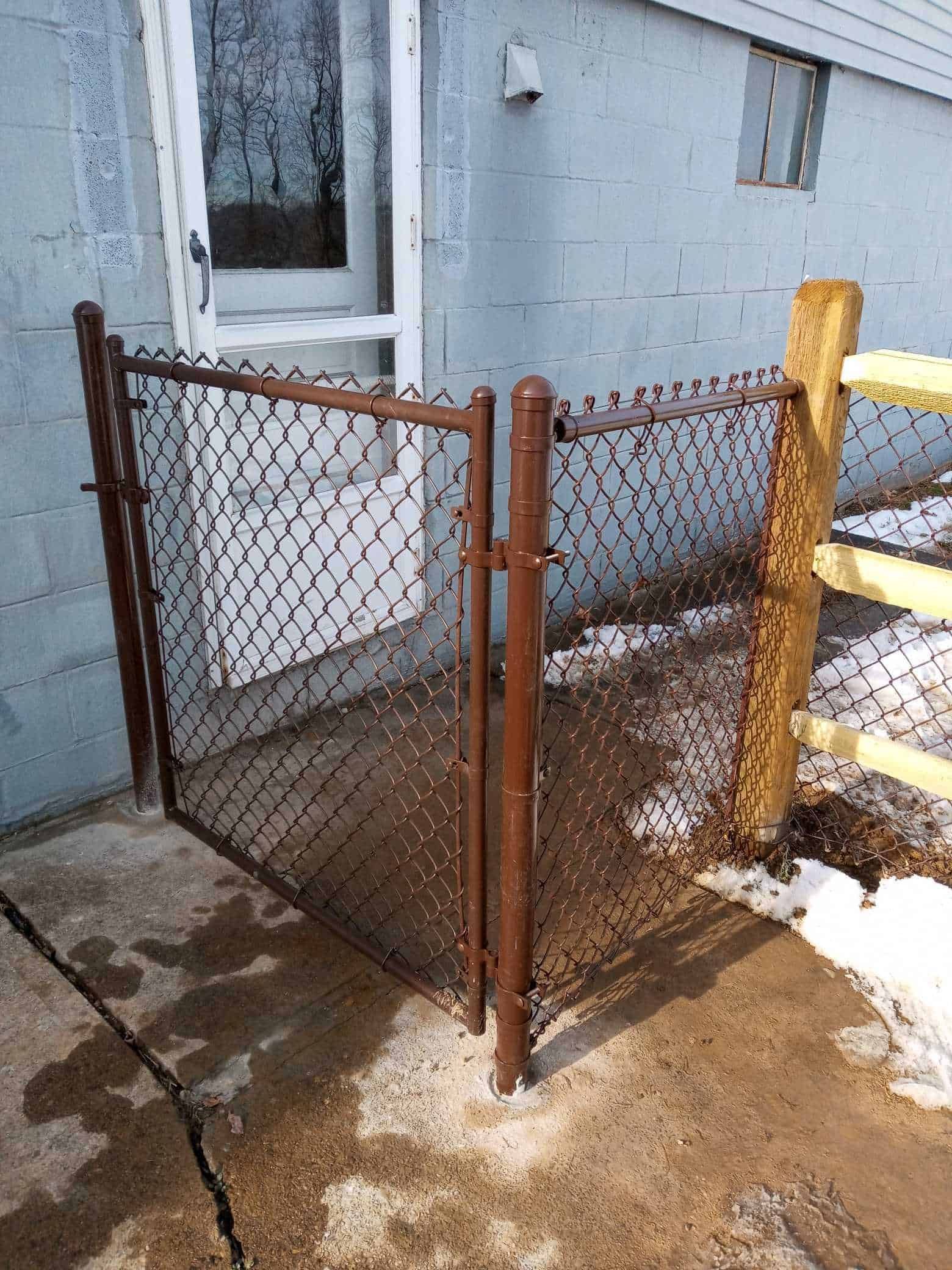 Chain-link fenced porch beside a blue house, with wet concrete and patches of snow.