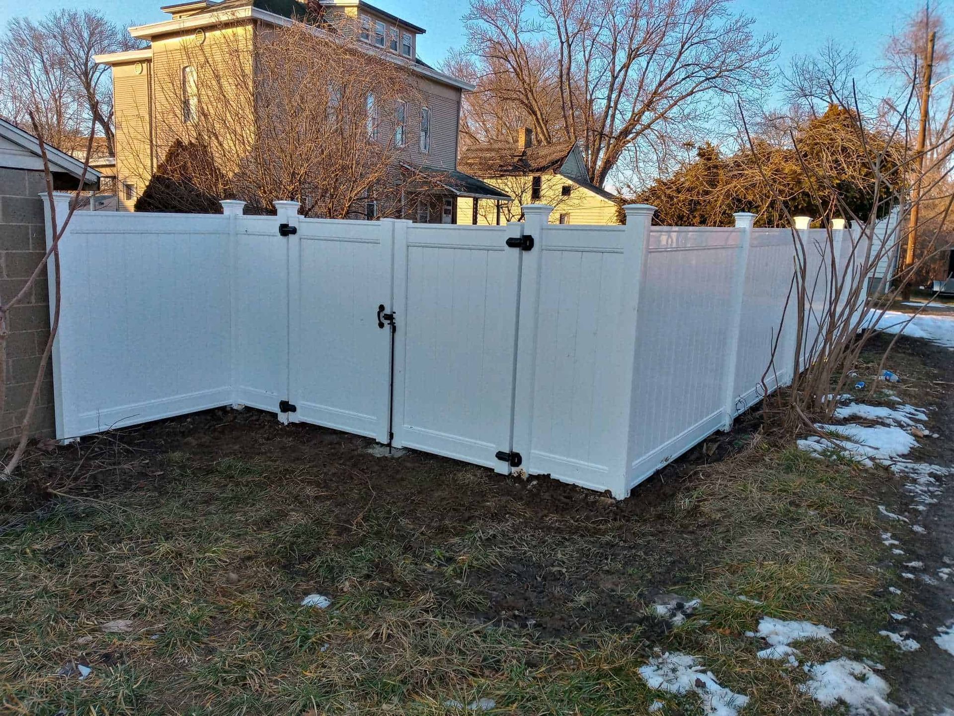 White vinyl fence enclosure in a yard with patches of snow and a stone house in the background