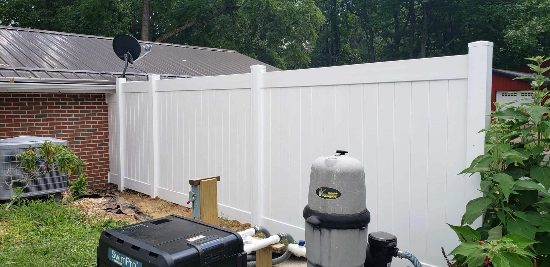 White vinyl privacy fence in a backyard beside a grill and patio equipment, with trees and brick house nearby