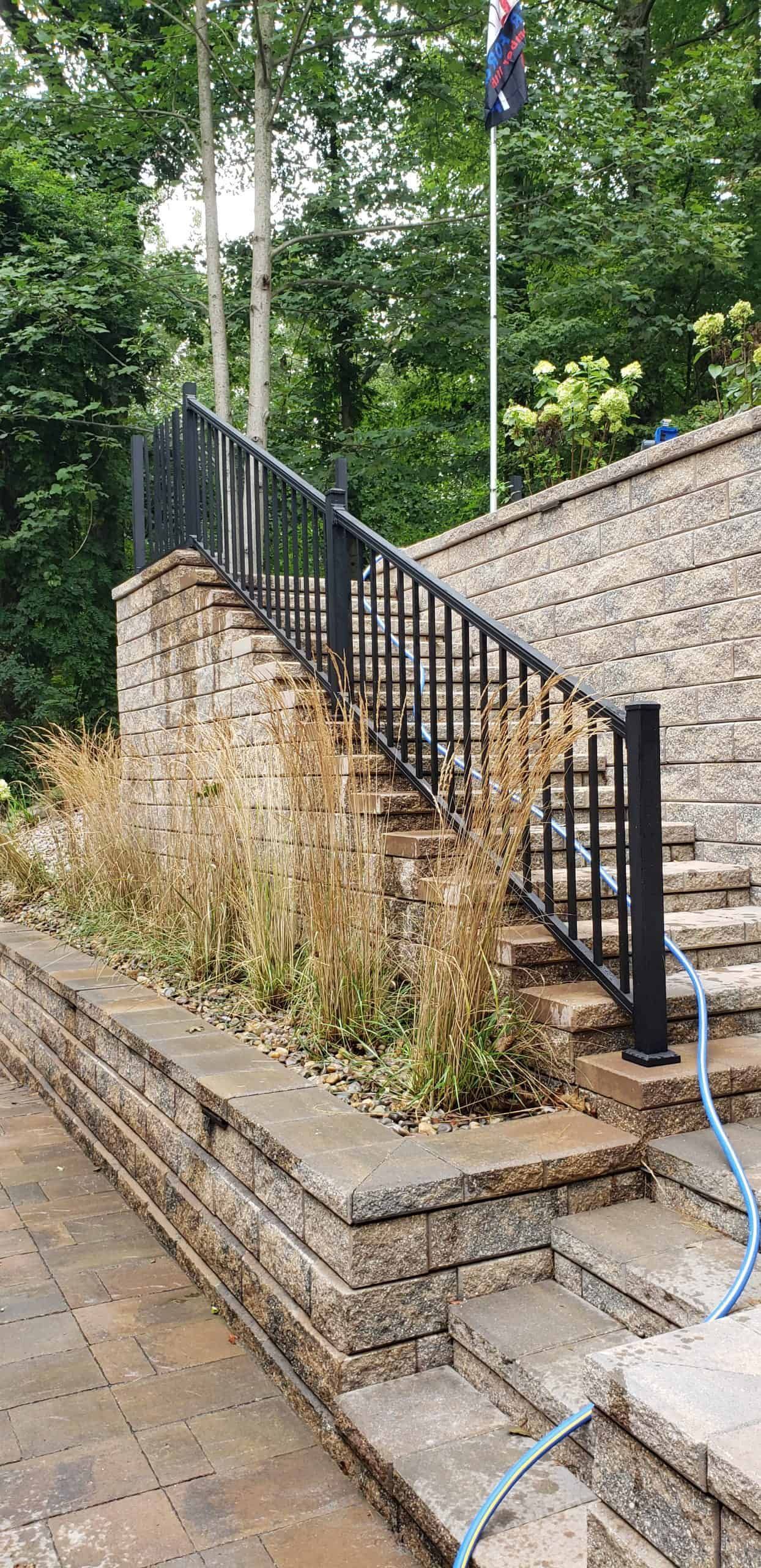 Brick stairway with black handrail beside a stone wall and overgrown weeds