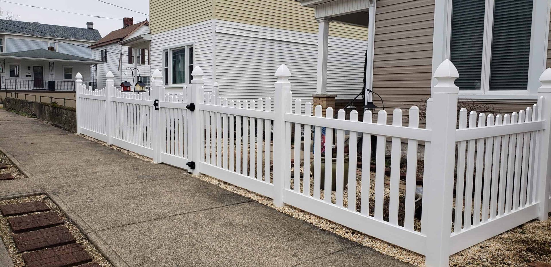 White picket fence along a sidewalk beside a white house with striped awning on a quiet street